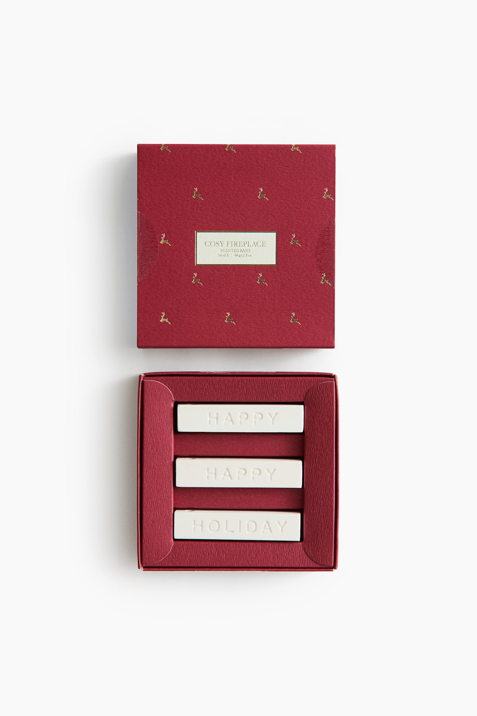 A scented bar gift set is presented in its open textured red box with the matching lid facing forward from a flat lay perspective. The lid features a scattered pattern of small gold-foiled reindeer and a central label, while the open compartment holds three white rectangular wax bars embossed with the words "HAPPY", "HAPPY", and "HOLIDAY".