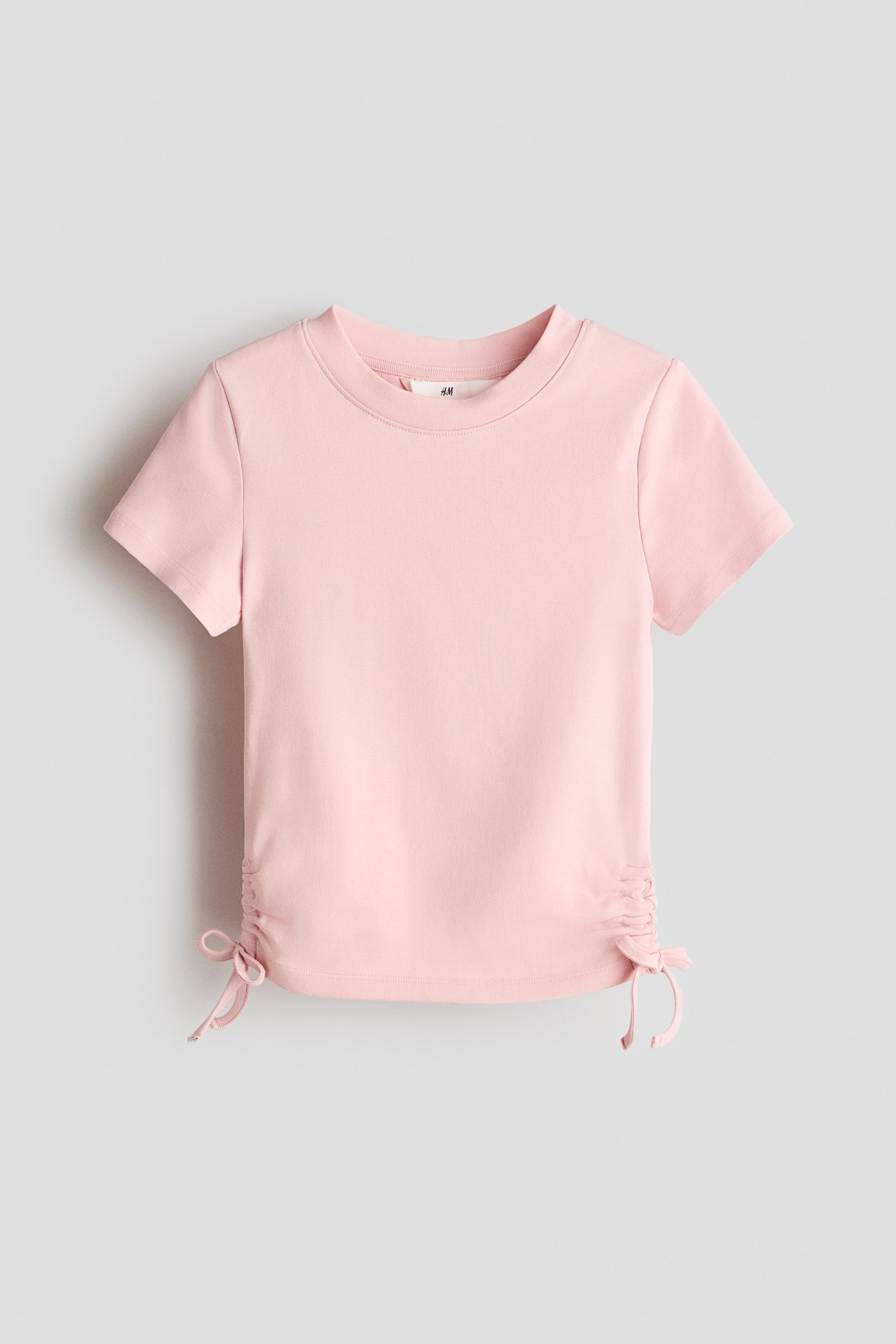 Xem ảnh lớn hơn: Áo thun ngắn tay màu hồng bằng chất liệu cotton jersey được giới thiệu với mặt trước, có cổ tròn. Dây rút trang trí với nơ may cố định tạo điểm nhấn, thu gọn vải ở hai bên gấu áo.