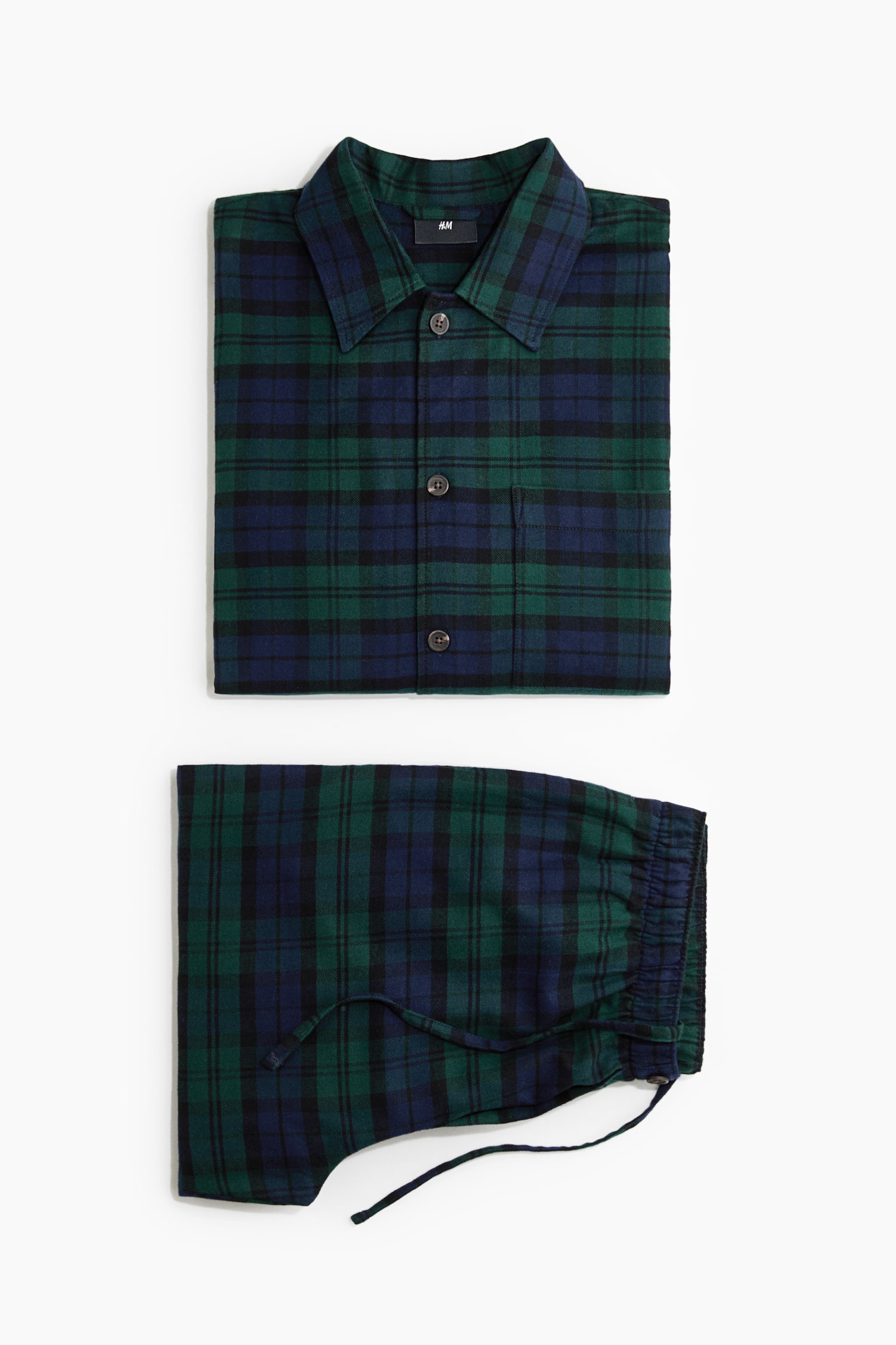 A green and navy blue plaid cotton flannel pyjama set is laid flat, showcasing a neatly folded button-up shirt with a turn-down collar and a single chest pocket, alongside matching folded pants with an elasticated waistband and drawstring.