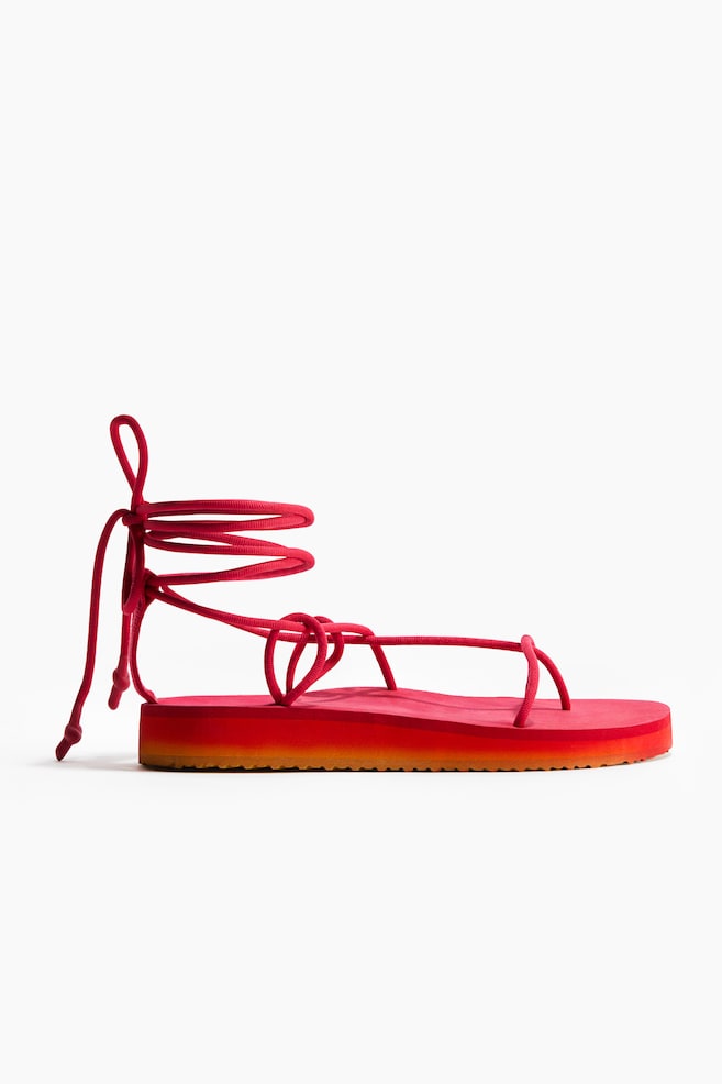Ein roter Schnürsandale wird aus einem seitlichen Winkel gezeigt, mit dünnen Kordelriemen, die eine Zehenschlaufe bilden und um den Knöchel wickeln, um sich zu einer Schleife zu binden. Die flache Plattformsohle zeigt eine rote Oberschicht über einer orangefarbenen Unterschicht. - 1