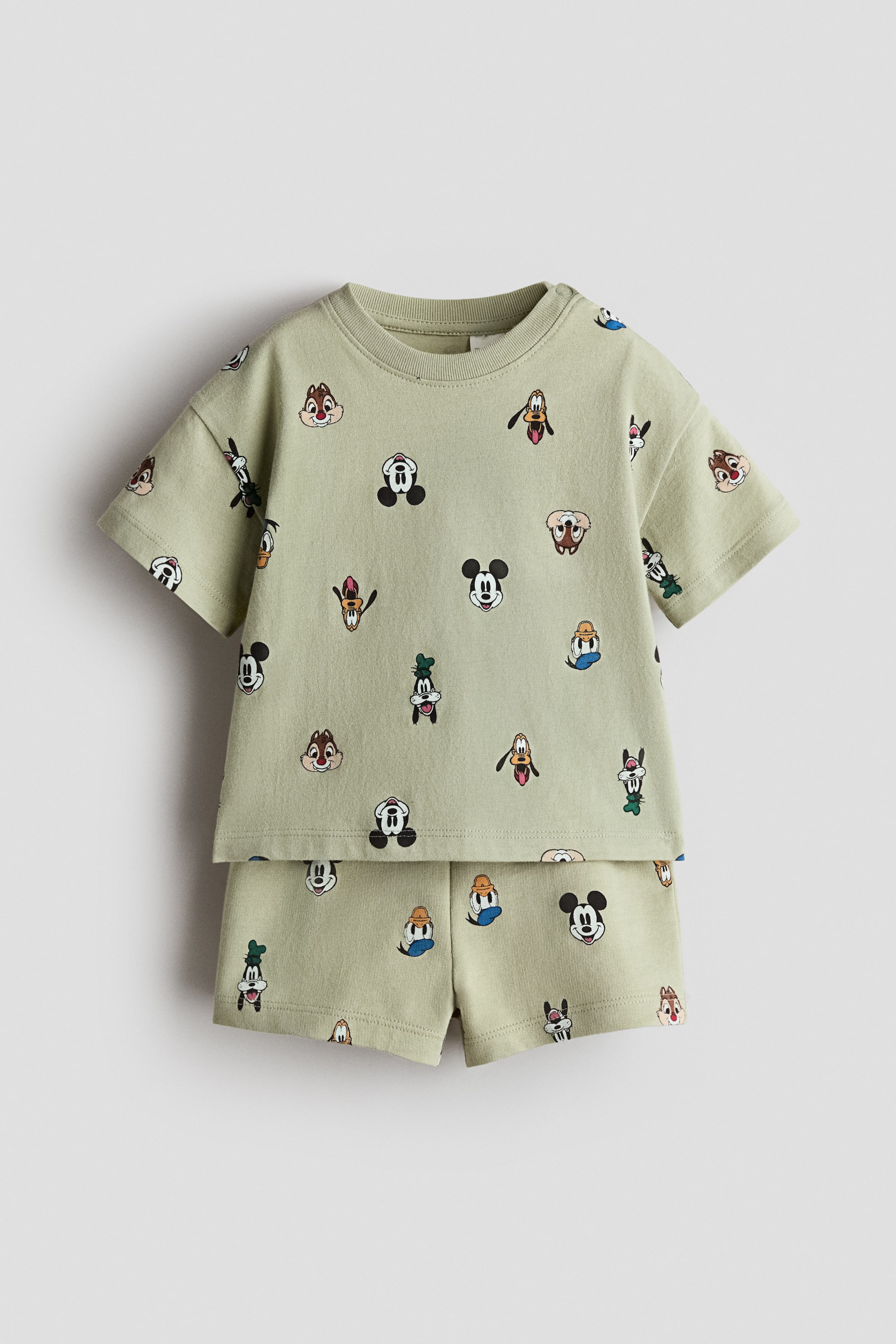 View larger image: A light green two-piece set is on display, comprising a short-sleeved t-shirt and shorts, both adorned with an allover print of various Disney character heads. The t-shirt features a ribbed crew neck and slightly overlaps the shorts, with the entire set laid flat and facing forward.
