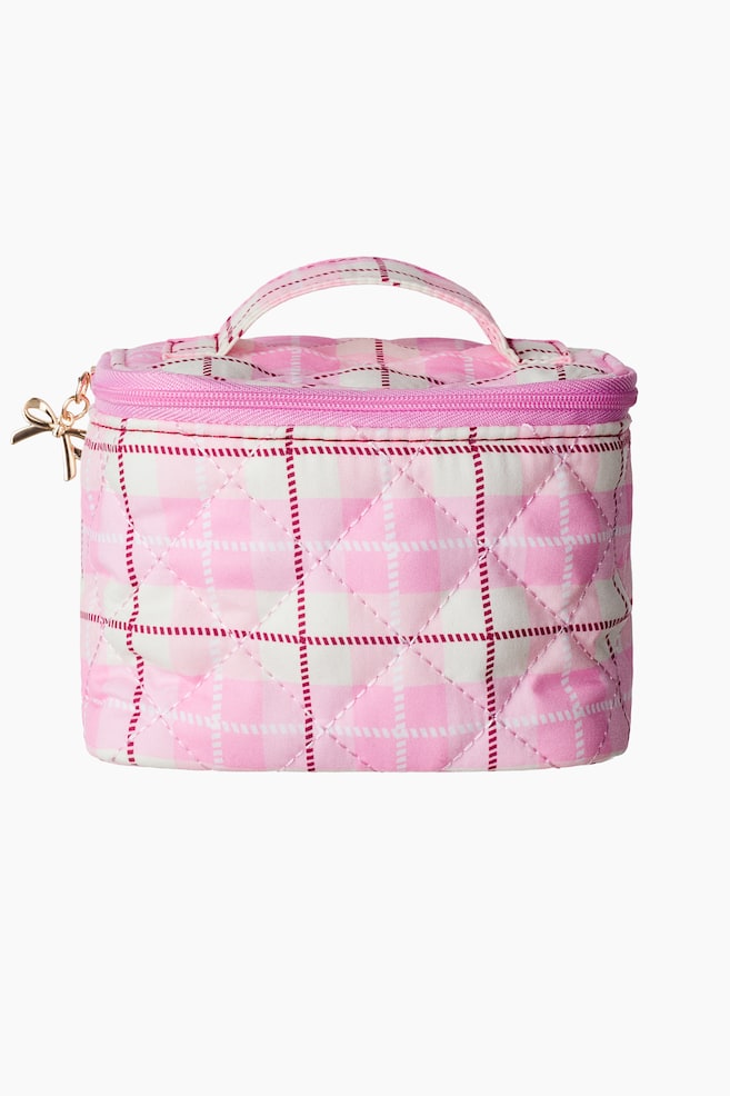 A pink and white quilted cosmetic bag is facing front, showcasing a plaid pattern with thin red dashed lines and a diamond-stitched texture. The bag is featuring a matching top handle, a pink zip-around closure, and a gold-tone bow-shaped zipper pull on the side. - 1