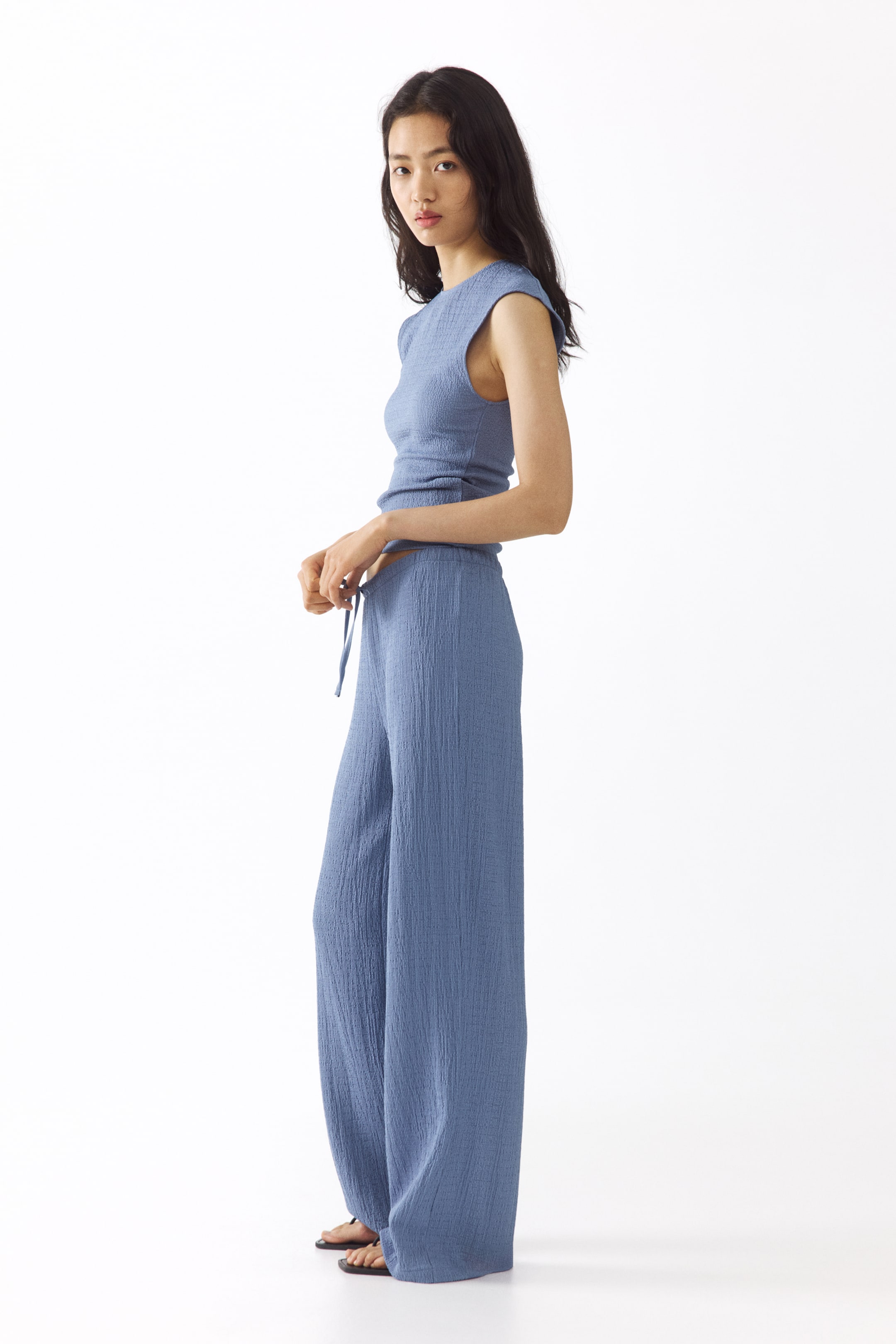 View larger image: The model is wearing blue textured jersey pants, designed with an elastic, drawstring waistband and straight legs. The model's hands are holding the drawstring.