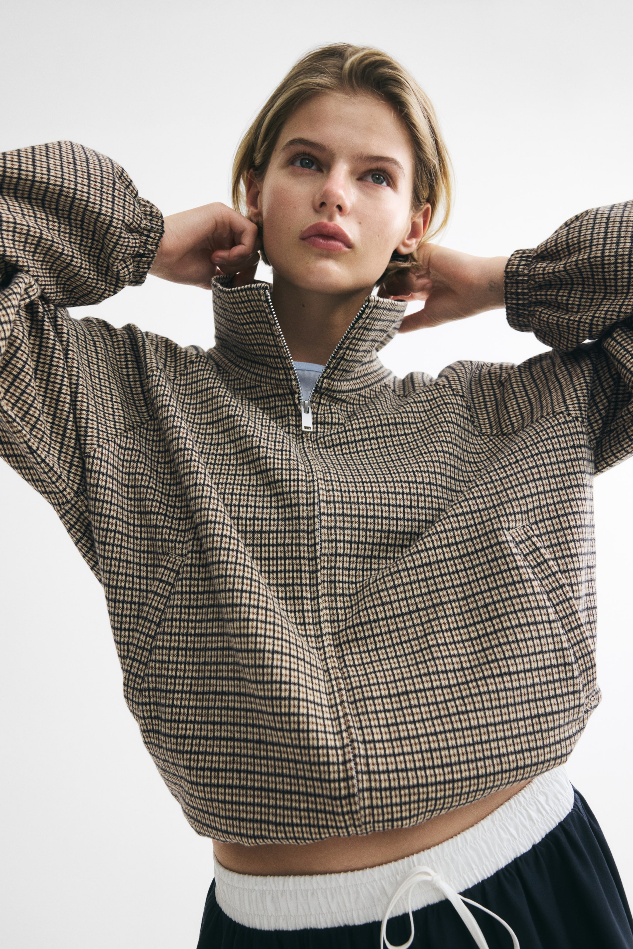 View larger image: A loose-fit jacquard knit jacket in a beige and dark check pattern is being worn by the model, who is raising their hands near its zipped funnel collar. The jacket's balloon sleeves are ending in elasticated cuffs, and a drawstring is cinching the hem for a puffy silhouette. It is being styled with dark trousers featuring a white elastic waistband and drawstring.