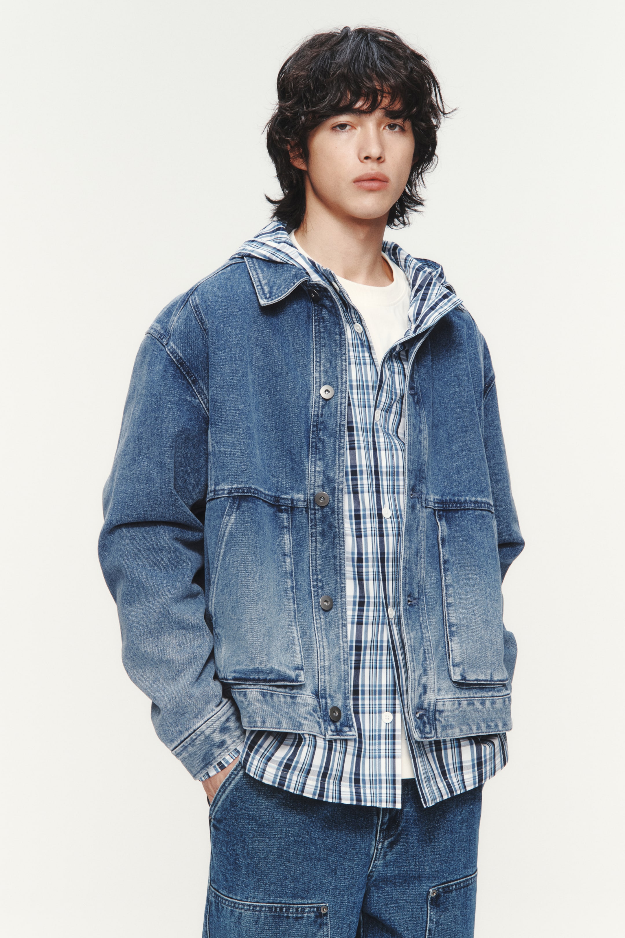 拡大画像を表示: ミディアムウォッシュのデニムジャケットは、クラシックなポイントカラーとボタンフロントクロージャーを備え、下部前面に2つの大きなパッチポケットがあります。ブルーとホワイトのチェックシャツとホワイトTシャツの上にオープンに着用され、リラックスしたフィット感を演出しています。