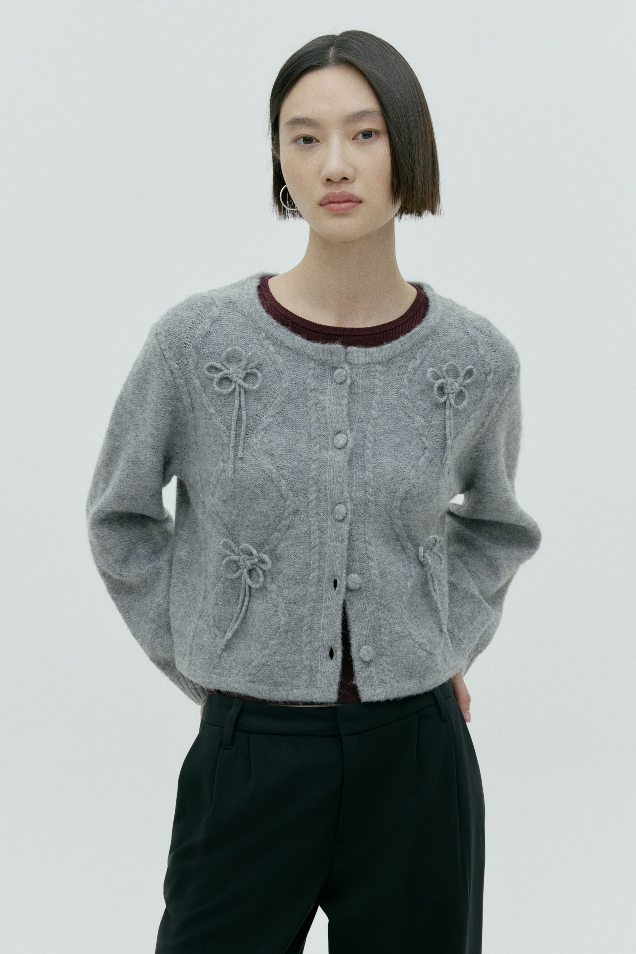 拡大画像を表示: グレーのケーブルニットカーディガンが正面やや斜めの角度で配置されており、前面にはリボン風の結び目の装飾があしらわれています。クルーネックで前開きのボタン仕様で、下部は開けた状態でマルーンのクルーネックトップスの上に羽織られています。