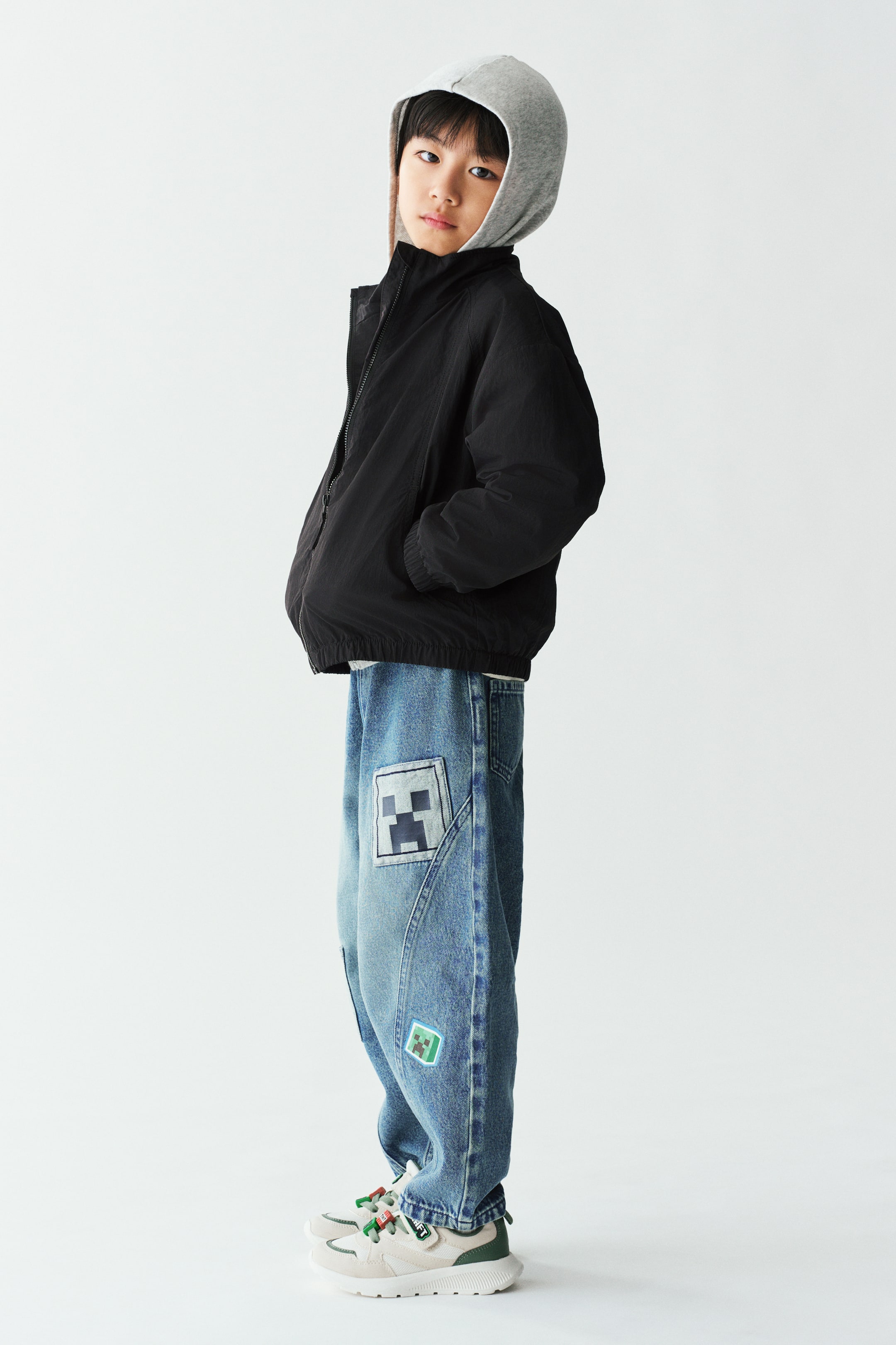 Xem ảnh lớn hơn: Quần jogger ống barrel bằng denim wash sáng đang được người mẫu mặc, nổi bật với phom dáng cong cùng các miếng patch hình vuông đồ họa ở bên hông ống quần. Quần jogger được phối cùng áo khoác đen khóa kéo, áo hoodie xám và giày sneaker trắng.