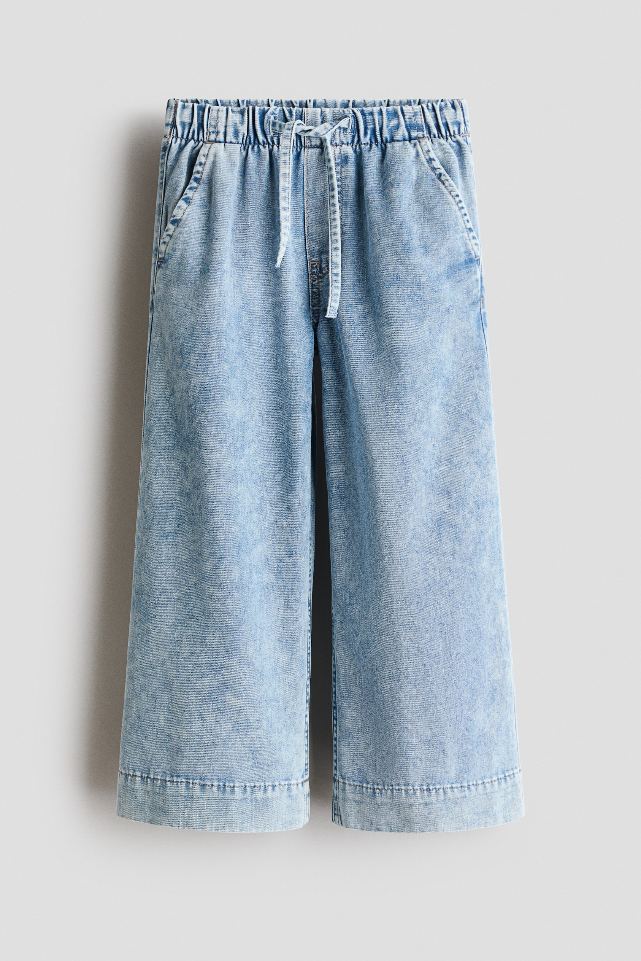 View larger image: Light blue denim culottes tergantung, menampilkan pandangan hadapan. Ia mempunyai pinggang berelastik berkerut dengan tali serut depan, dua poket sisi serong, dan kaki lebar yang dipotong dengan jahitan kelim.