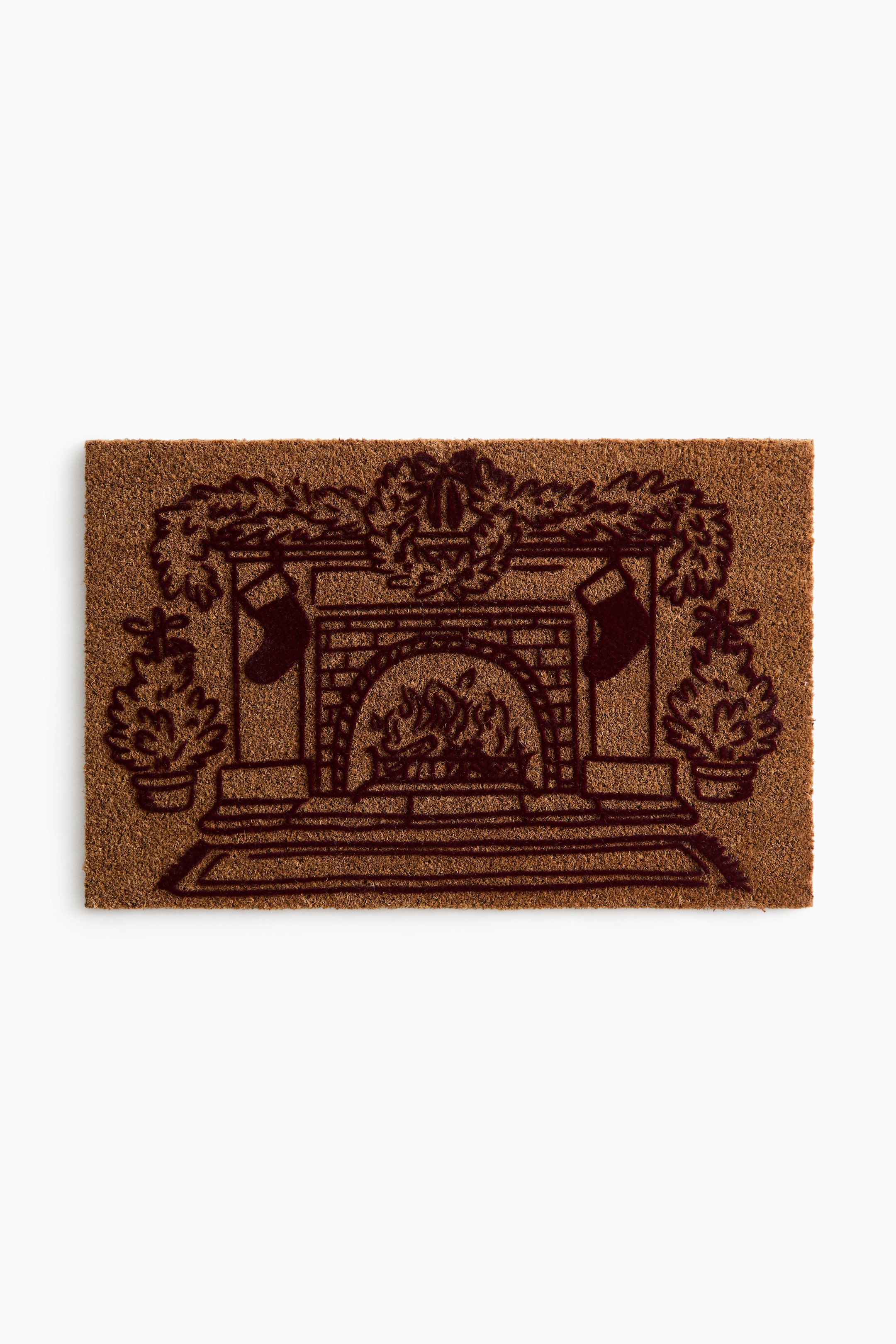 View larger image: A rectangular coir doormat features a dark brown printed illustration of a cosy fireplace scene. The design depicts a brick fireplace with a burning fire, two hanging stockings, a garland draped across the mantel, and potted evergreen trees flanking either side.