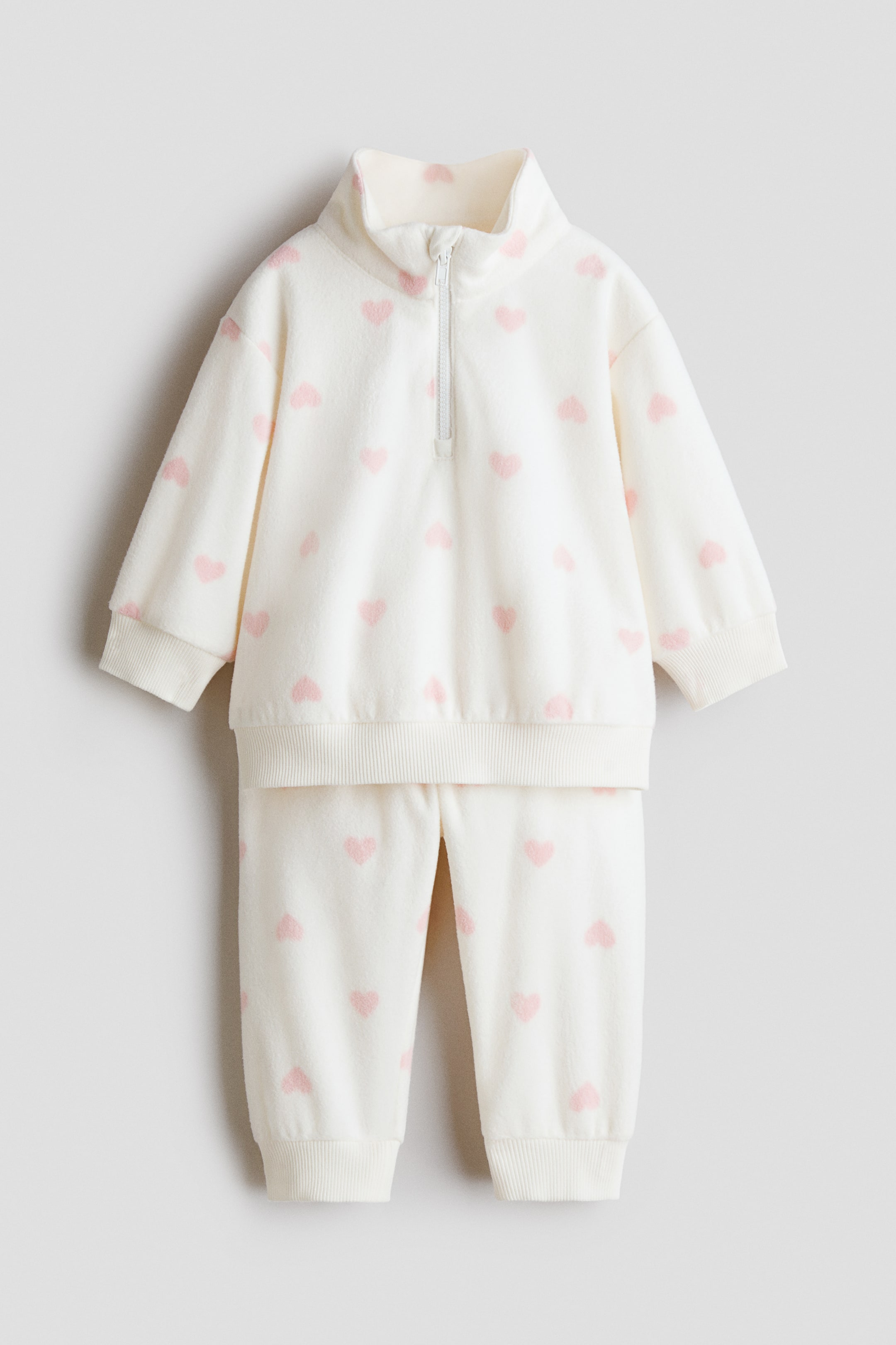 View larger image: A light cream two-piece set, shown front-facing, featuring a long-sleeved top and matching trousers, both with a pattern of pink hearts. The top is designed with a stand-up collar, a white half-zip closure, ribbed cuffs, and a ribbed hem, while the trousers feature ribbed ankle cuffs.