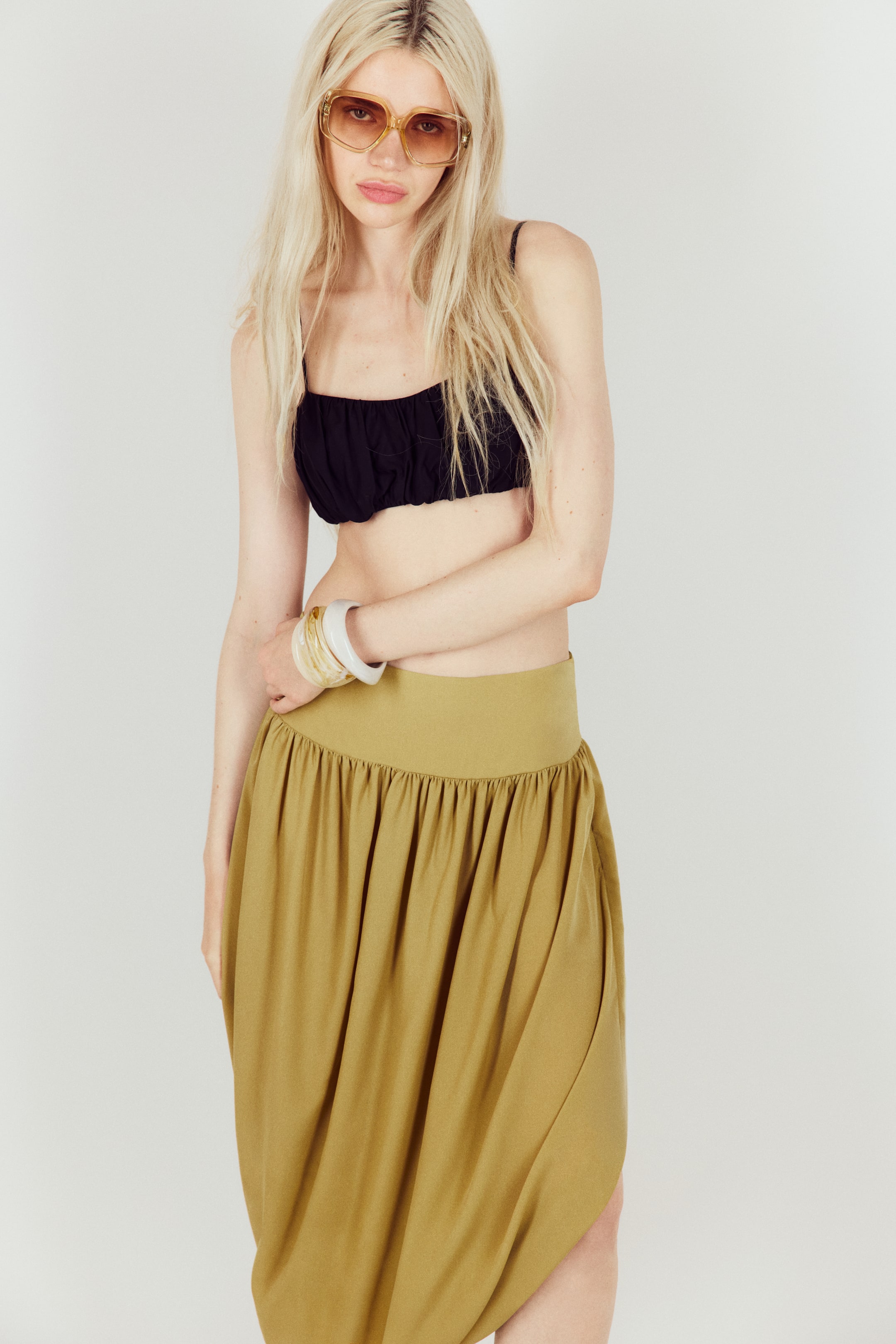View larger image: The model is wearing an olive green calf-length tulip skirt made from airy chiffon. It has a wide, gathered waistband and a bubble hem, giving it a puffy silhouette. The model's hand rests on the skirt's waistband.
