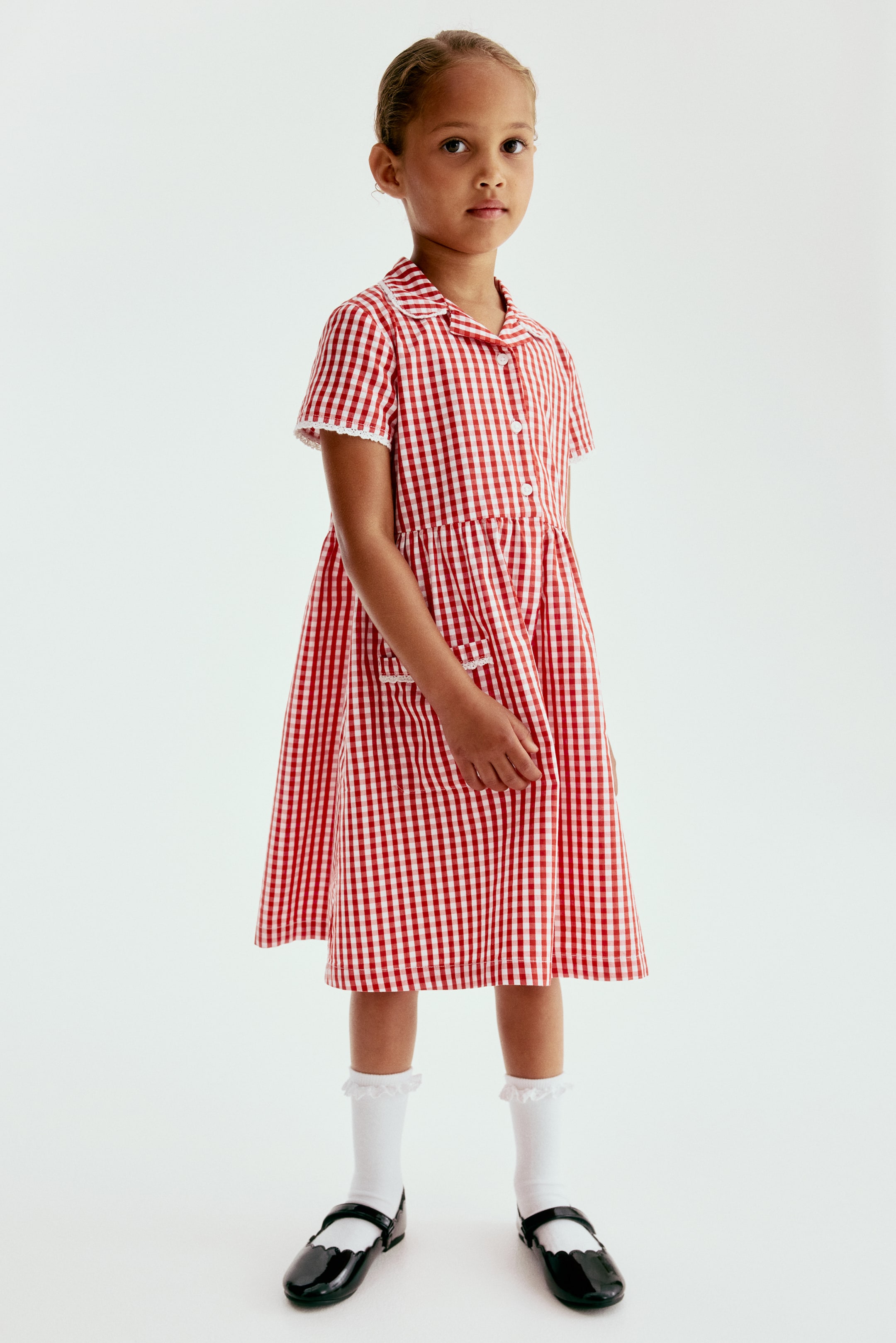 View larger image: A school dress featuring a red and white checked cotton weave, a rounded collar, a button placket with white buttons, and short sleeves. It is designed with a gathered seam at the waist, a flared skirt with patch front pockets, and lace trim detailing the collar, sleeve cuffs, and pocket openings.