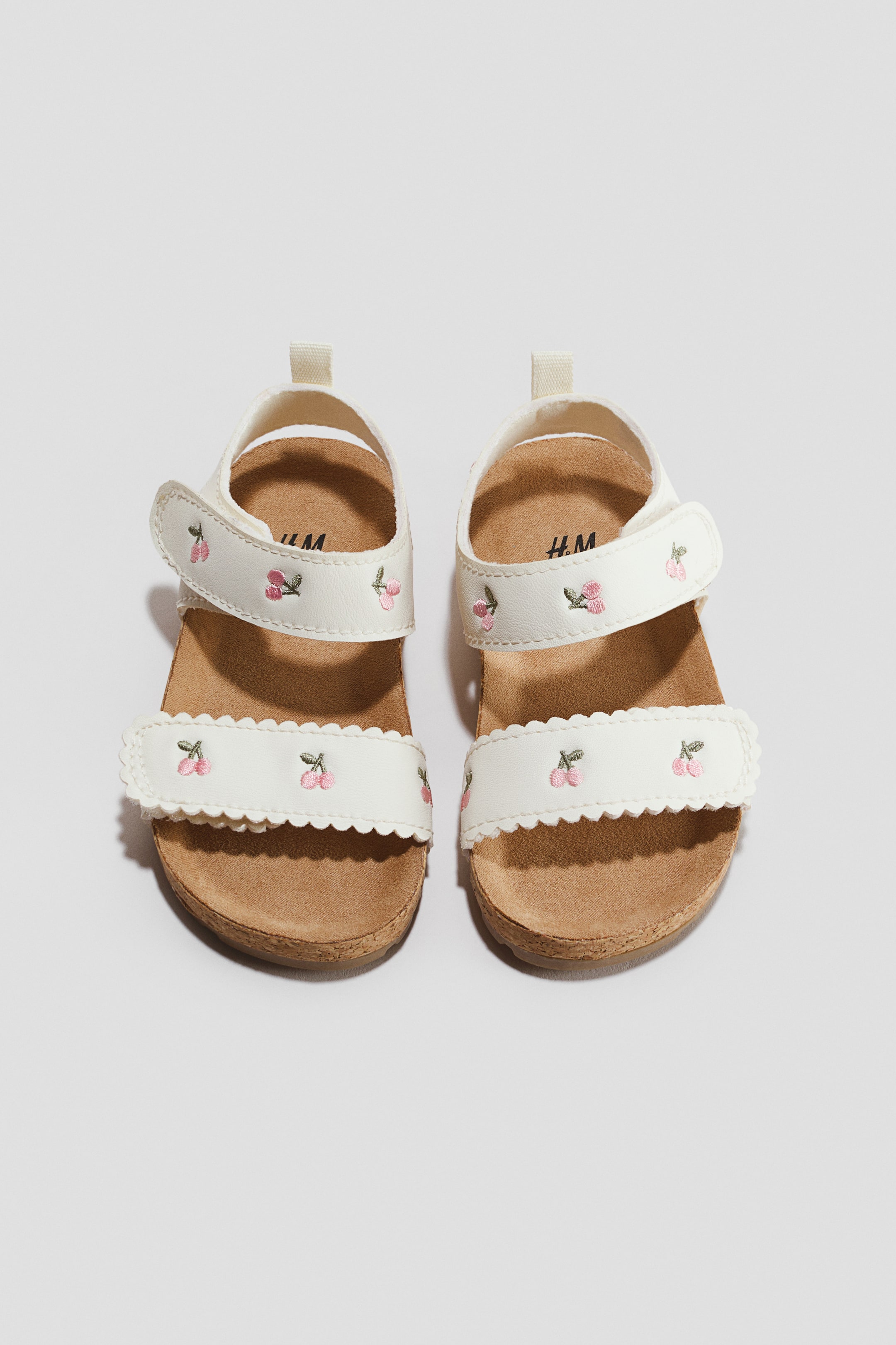 View larger image: A pair of sandals is facing forward, featuring white straps decorated with small embroidered pink cherries. The design includes a scalloped foot strap, an adjustable ankle strap with a hook-and-loop fastener, a white heel pull tab, and contoured brown moulded insoles.