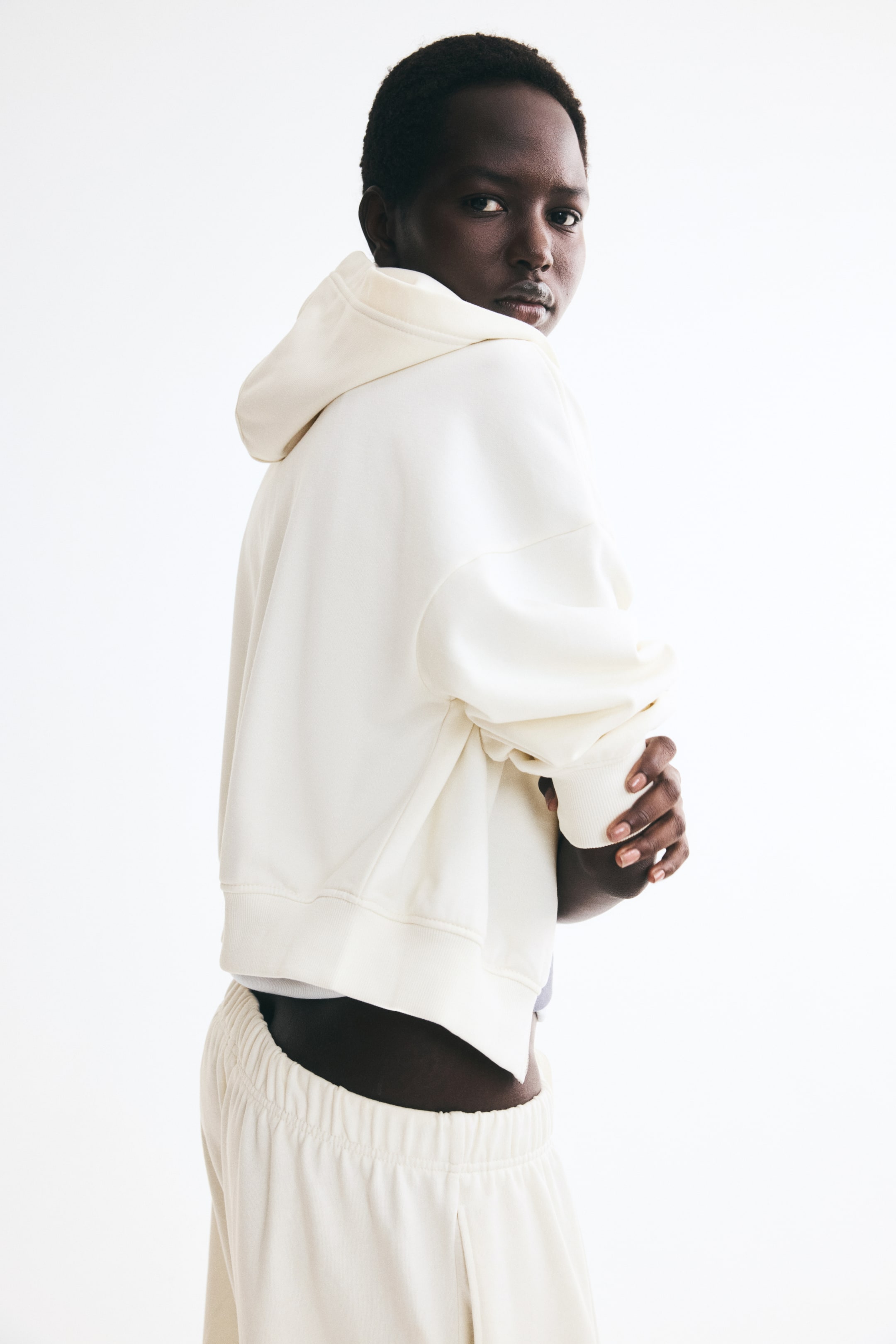 View larger image: A cream-colored cropped hoodie is being worn by the model, who is standing with her arms crossed to showcase the back and side profile of the garment. The lightweight sweatshirt-style hoodie features a double-layered hood, dropped shoulders, and ribbed cuffs and hem, styled with matching cream sweatpants.