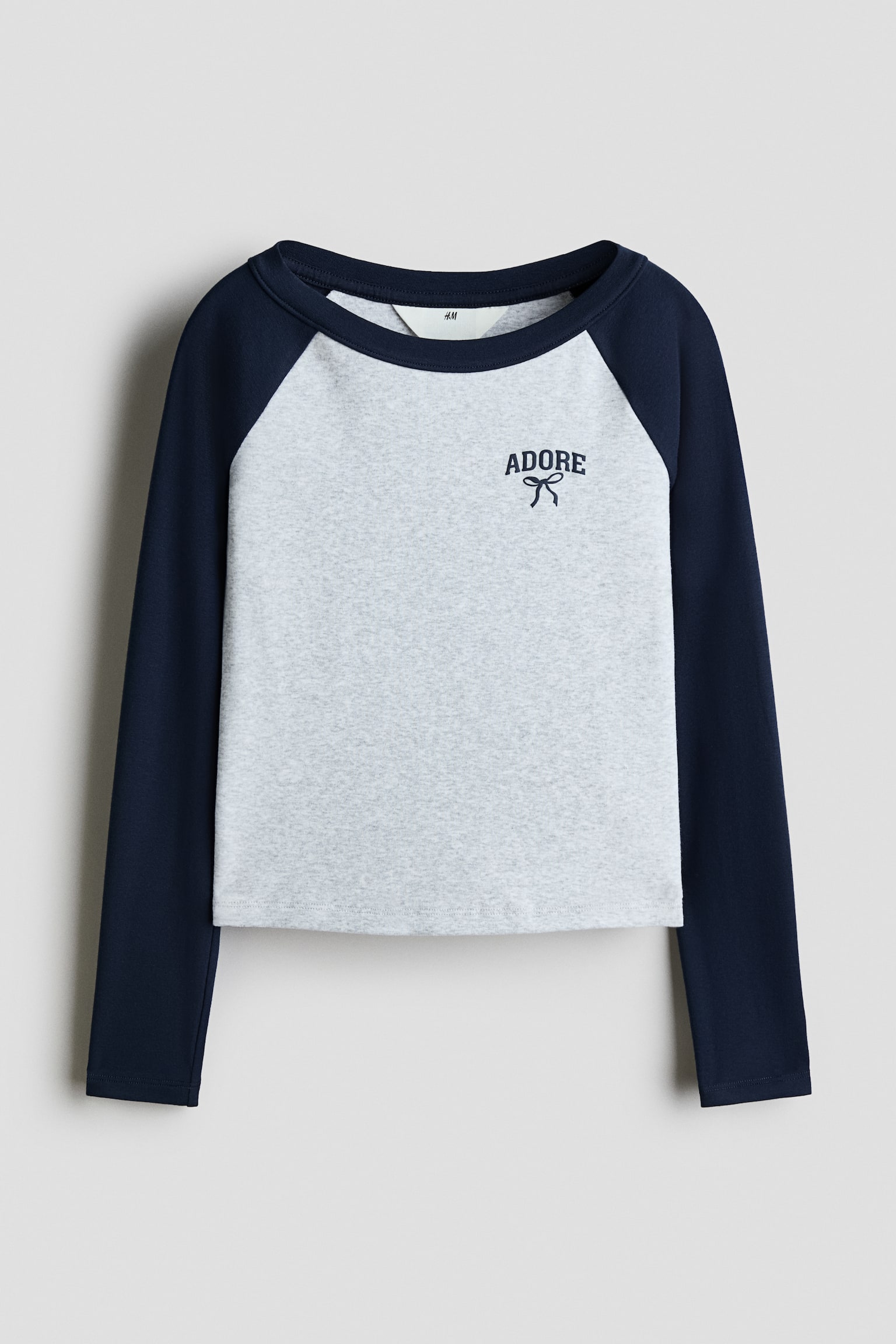 Dit frontale raglan-T-shirt met lange mouwen toont een lichtgrijs gemÃªleerd lichaam met contrasterende marineblauwe mouwen en een bijpassende ronde hals. Op de linkerborst bovenaan staat een kleine marineblauwe graphic met het woord 'ADORE' boven een eenvoudige strikillustratie.