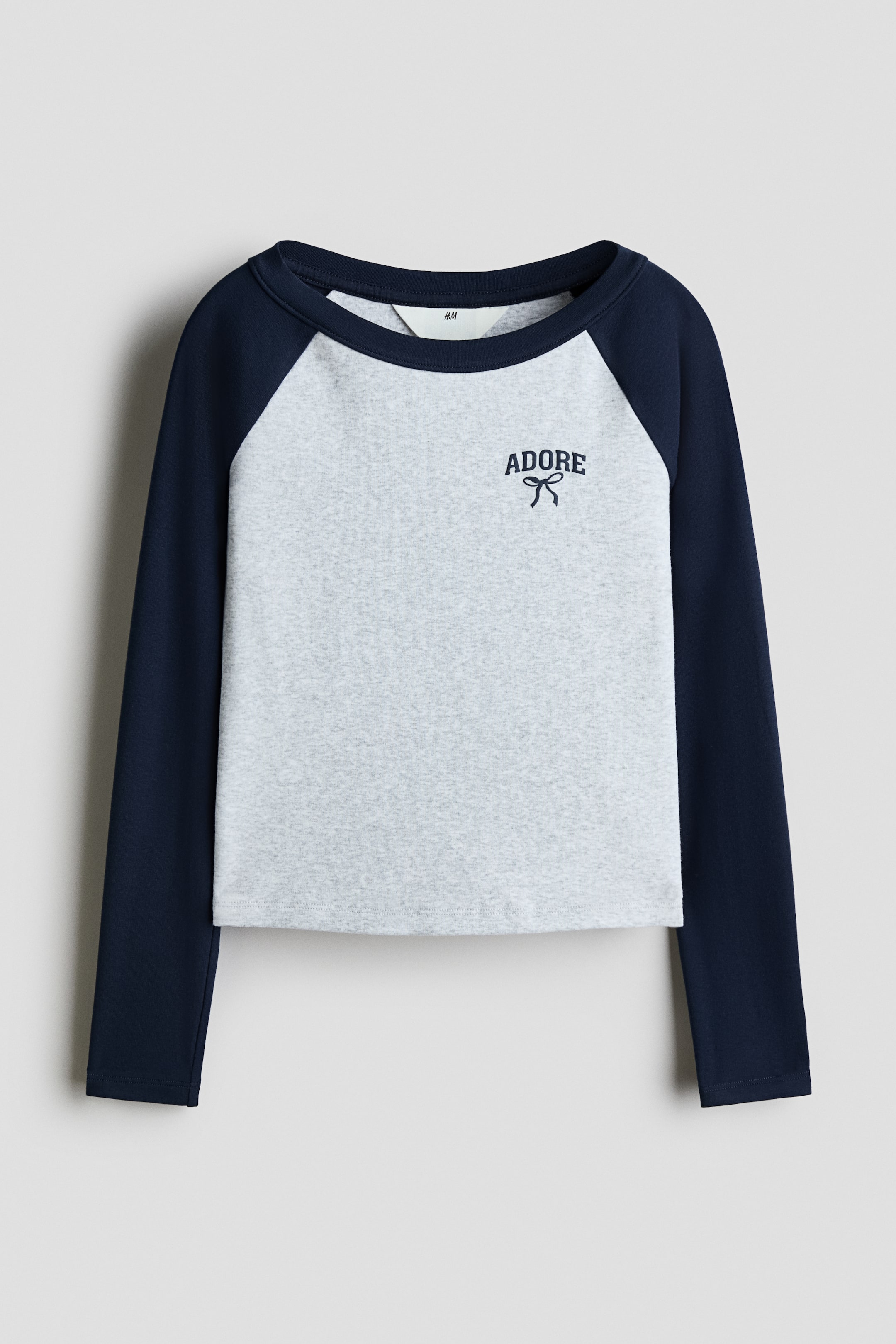 View larger image: This front-facing, long-sleeve raglan T-shirt features a light gray heather body with contrasting navy blue sleeves and a matching crew neckline. A small navy blue graphic displaying the word "ADORE" above a simple bow illustration is shown on the upper left chest.