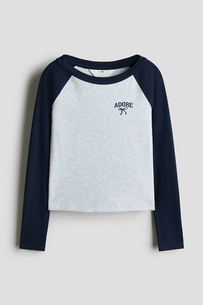 Ten ujÄty od przodu T-shirt z dÅugim rÄkawem i raglanowymi rÄkawami prezentuje jasnoszare, melanÅ¼owe body z kontrastowymi granatowymi rÄkawami oraz dopasowanym okrÄgÅym dekoltem. Na gÃ³rnej lewej czÄÅci klatki piersiowej znajduje siÄ niewielki granatowy nadruk z napisem âADOREâ umieszczonym nad prostÄ ilustracjÄ kokardy. - 1
