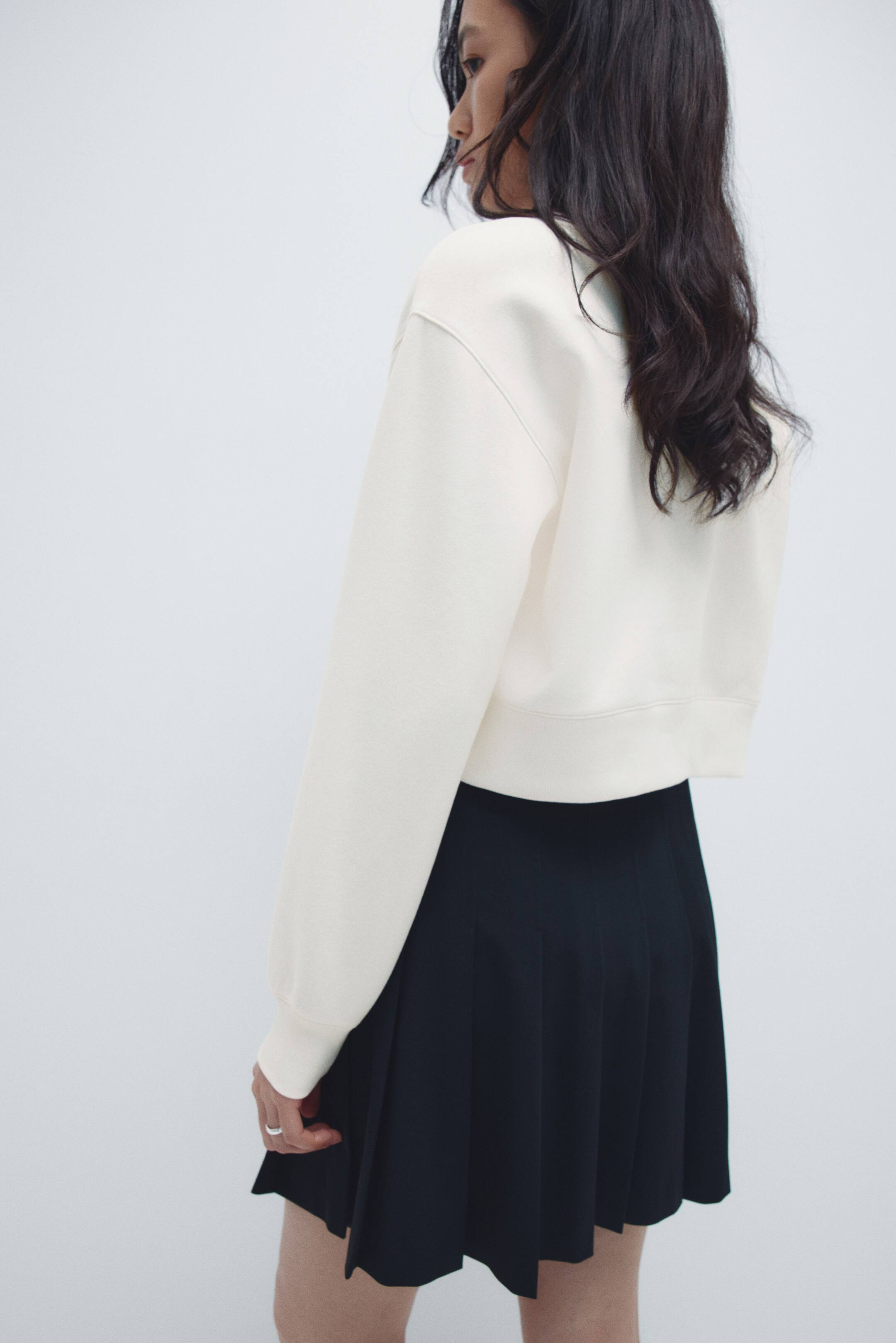 이미지 확대 보기: 크림색 크롭 스웨트셔츠는 긴 소매와 리브드 커프, 그리고 리브드 밑단이 특징이며, 세로 쳐다본 3/4 각도에서 보이고 있습니다. 이는 검정 주름 미니 스커트와 함께 착용되어 있으며 스커트가 퍼지고 있습니다.