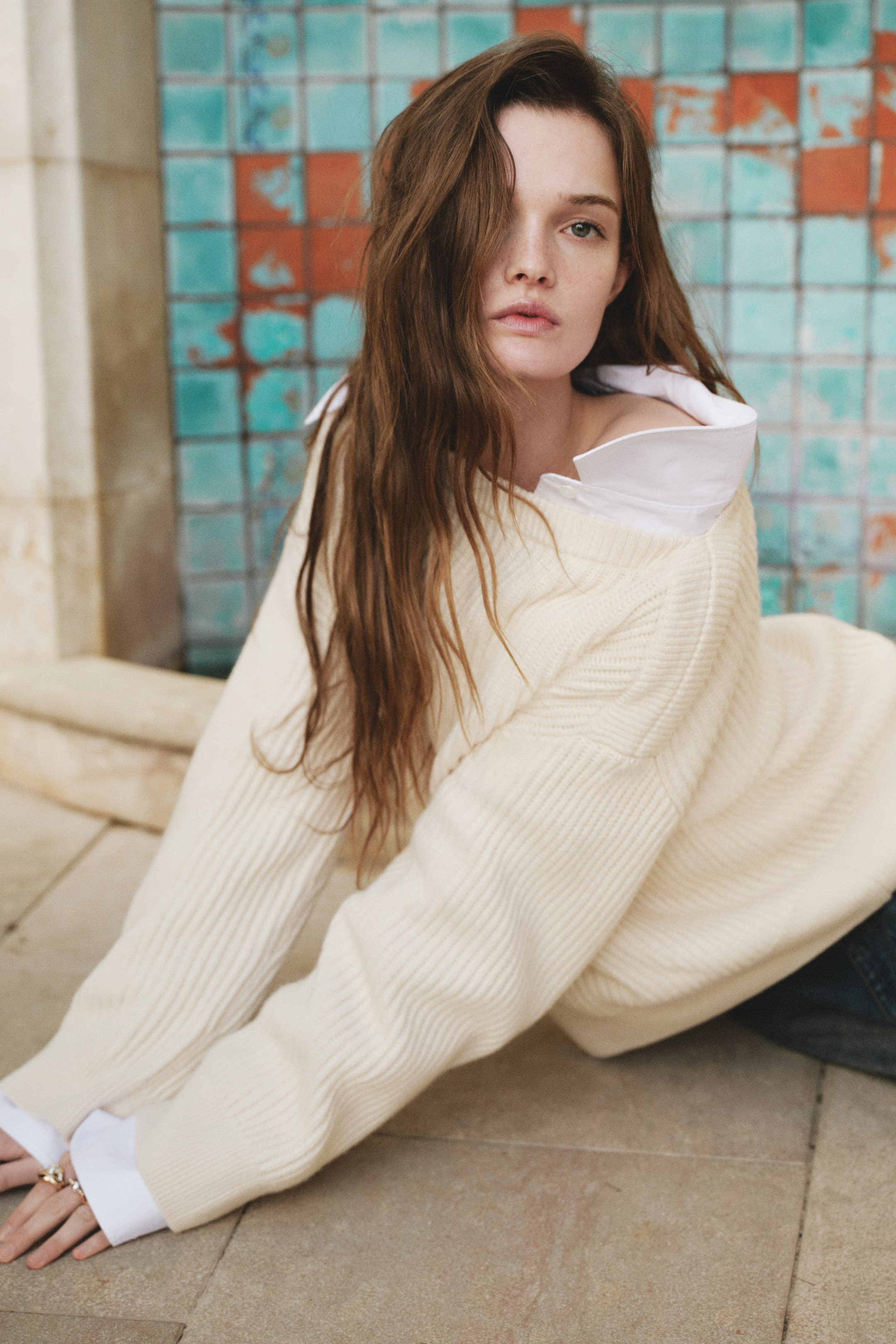 Visualizza immagine più grande: Un maglione a maglia color crema presenta una texture a coste prominente sulle maniche lunghe, che terminano con polsini a coste, e un sottile motivo a maglia diagonale sulla parte superiore del braccio. Viene indossato in uno stile rilassato e sceso sulle spalle sopra una camicia bianca con colletto, con il colletto e i polsini della camicia visibili.