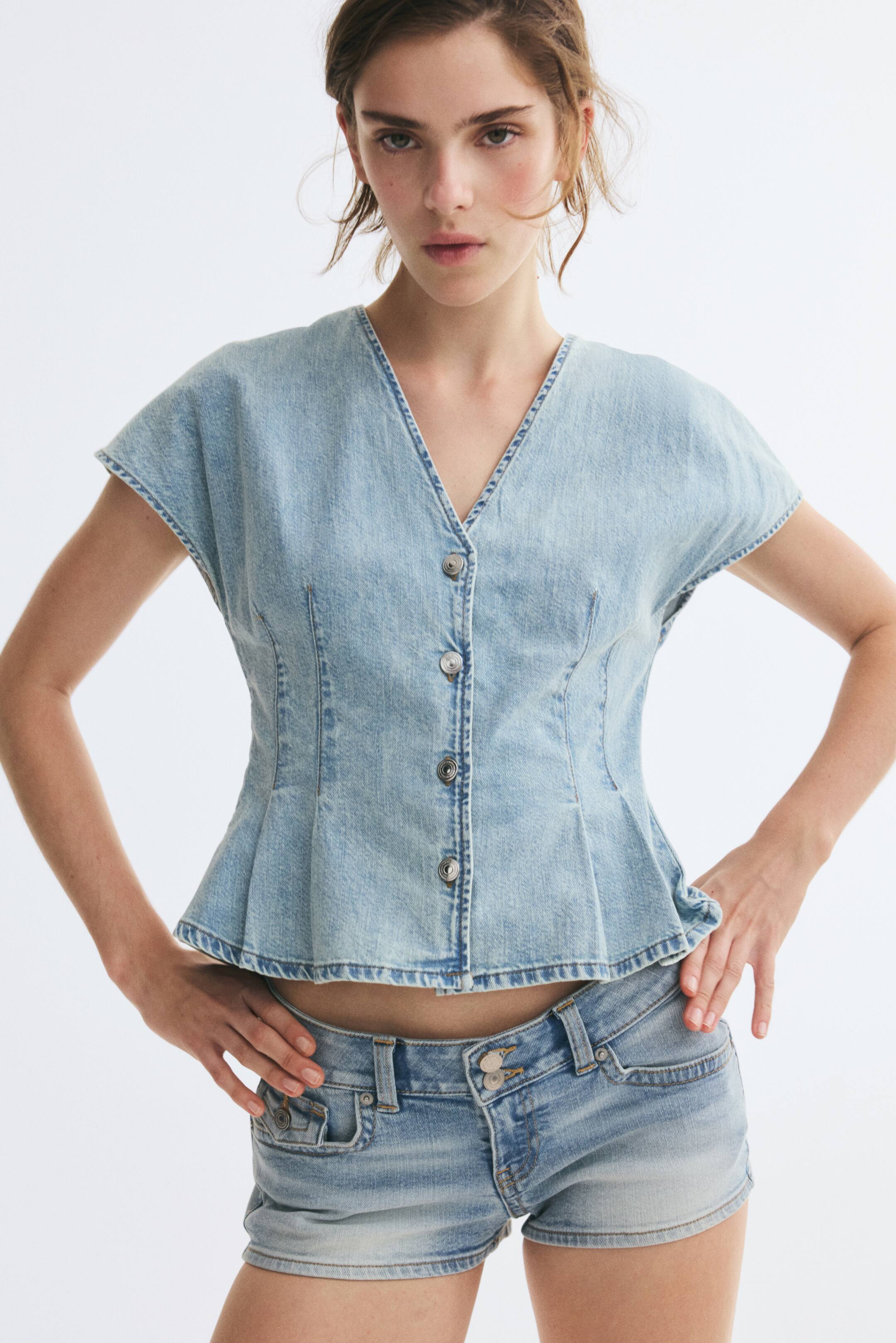 View larger image: The model is wearing a light-wash blue denim top featuring a V-shaped neckline, button-up front, cap sleeves, and a flared peplum hem created by waist darts. The top is paired with matching denim shorts, and the model is standing with hands resting on the hips.