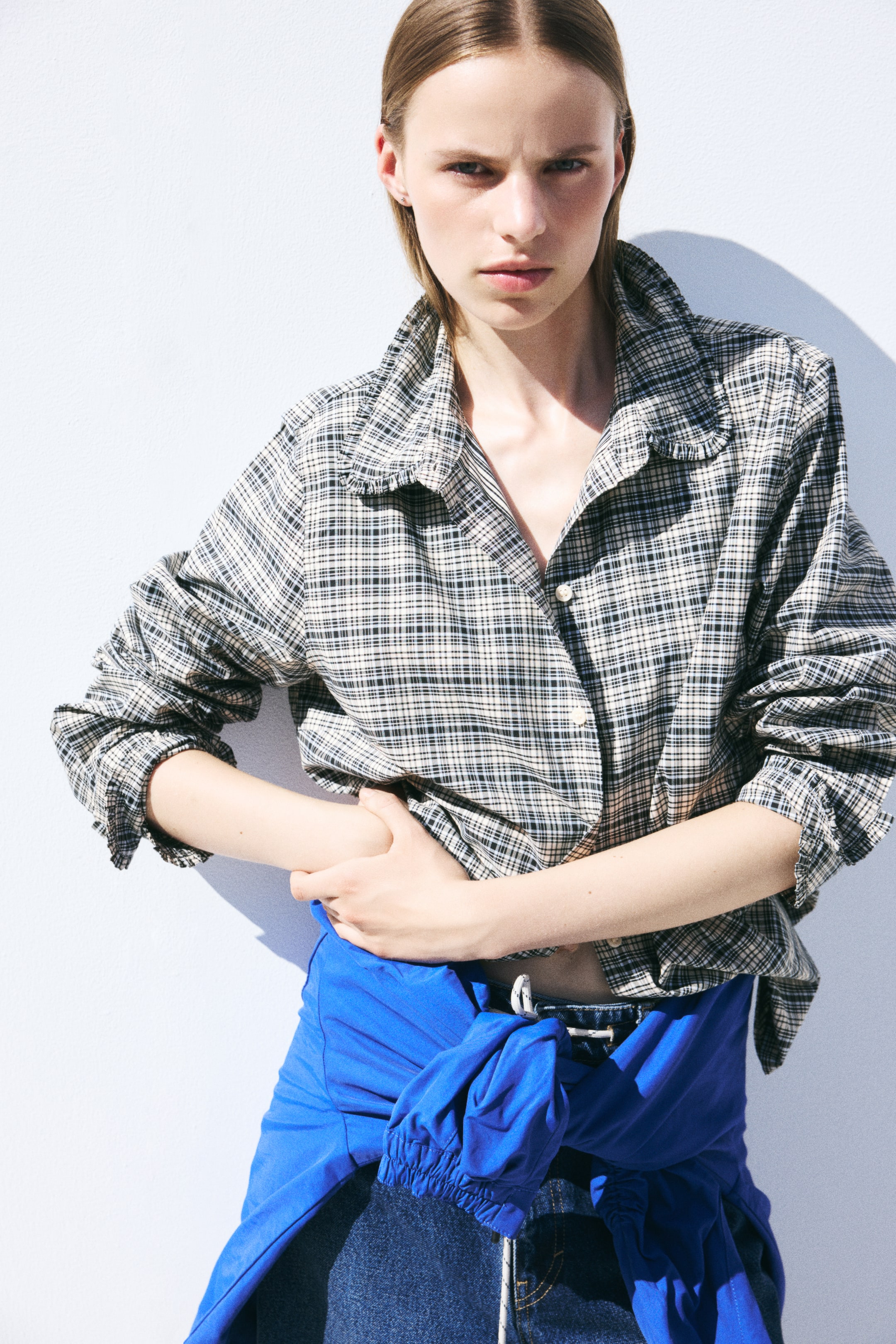 Visualizza l'immagine ingrandita: La modella indossa una camicia a quadri a maniche lunghe con colletto rifinito da volant, con i bottoni anteriori parzialmente slacciati e le maniche arrotolate. La camicia è abbinata a jeans in denim scuro e a un capo di colore blu acceso annodato in vita.