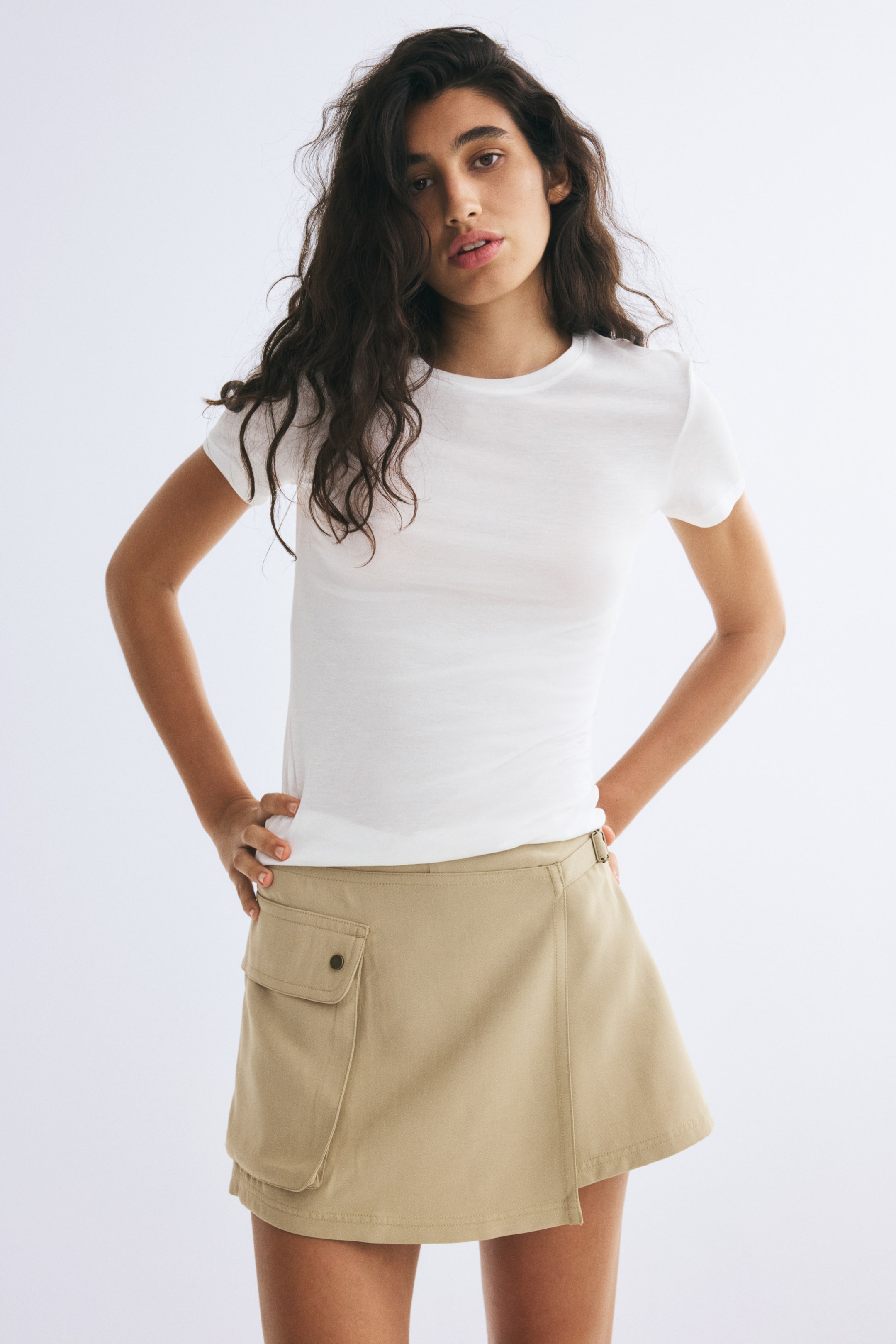 View larger image: A beige short skort is being worn by the model, styled with a fitted white T-shirt. The high-waisted skort features a sewn-in wrap front with a side buckle and a large bellows pocket with a flap on the right side.