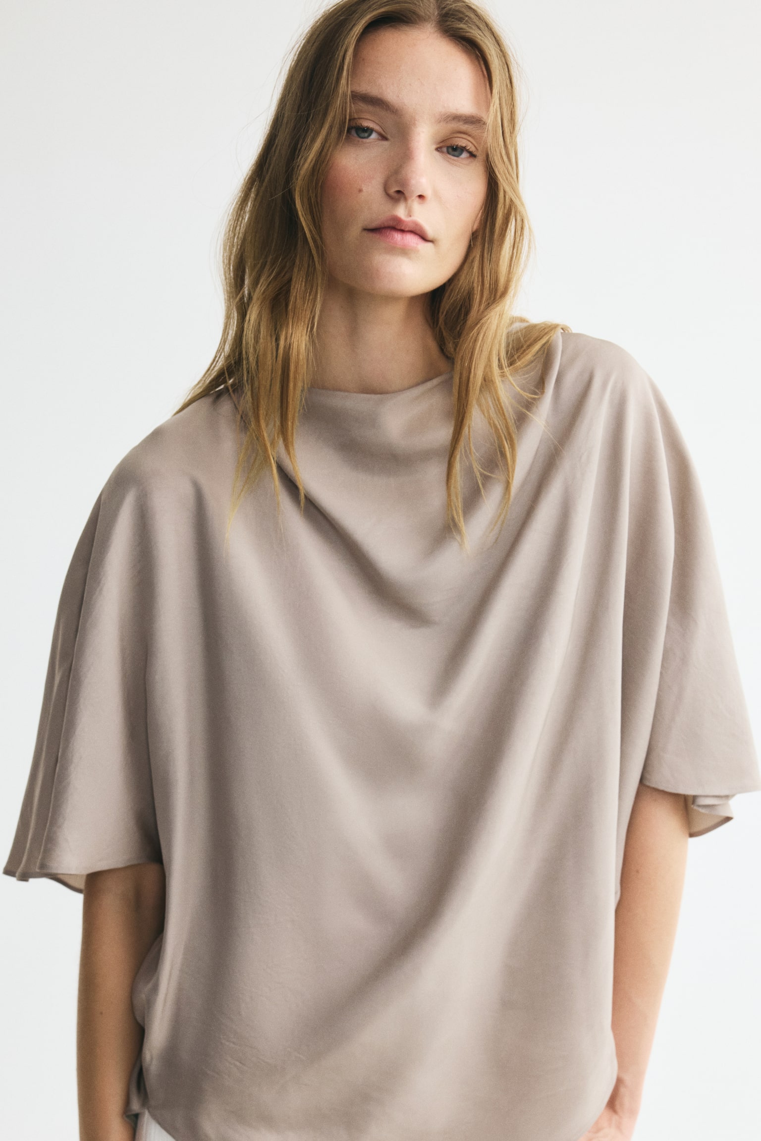 A taupe loose-fitting top is showing its back, featuring a draped neckline, wide short sleeves, and a rounded hem. - 1