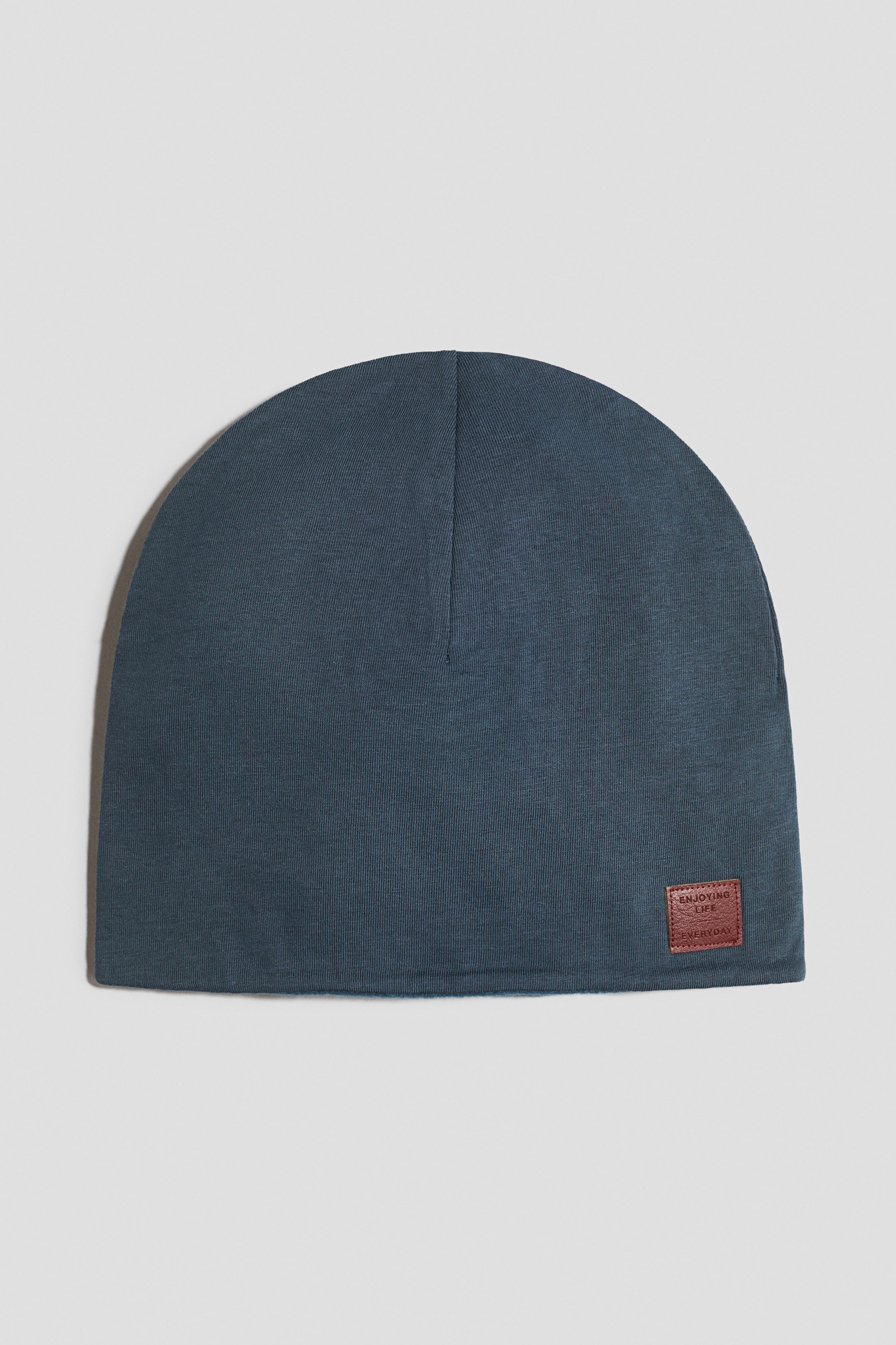 View larger image: A dark teal beanie is shown from the front, featuring a soft, rounded crown. A small, rectangular, reddish-brown patch is stitched onto the lower right side, displaying embossed text.