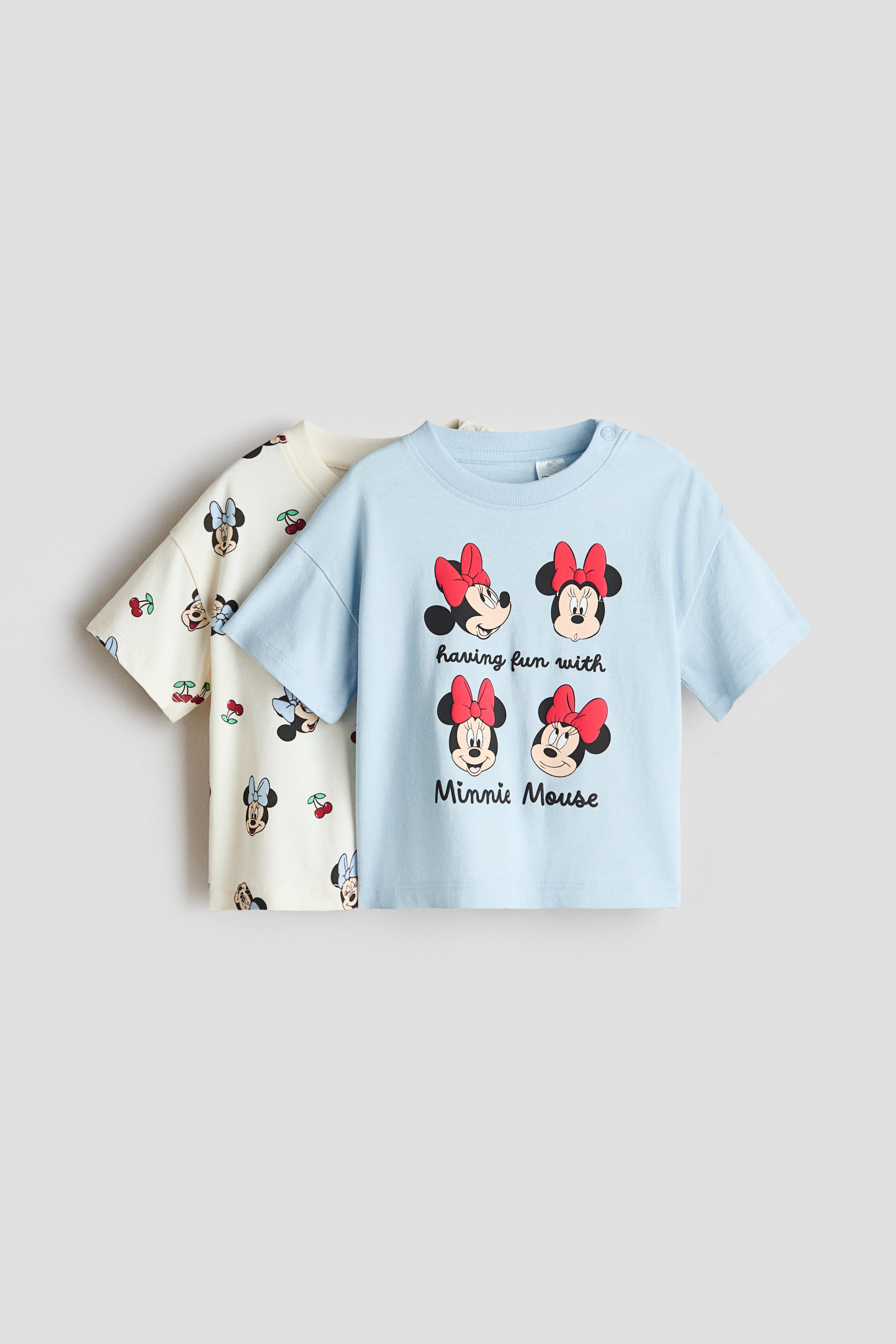 View larger image: A 2-pack of printed cotton T-shirts is showcasing a light blue T-shirt at the front, adorned with four different Minnie Mouse head illustrations featuring red bows and the phrase 'having fun with Minnie Mouse' on the front. It includes snap buttons on the left shoulder for added convenience. Behind it, an off-white T-shirt displays an all-over print of Minnie Mouse heads with light blue bows and red cherries.