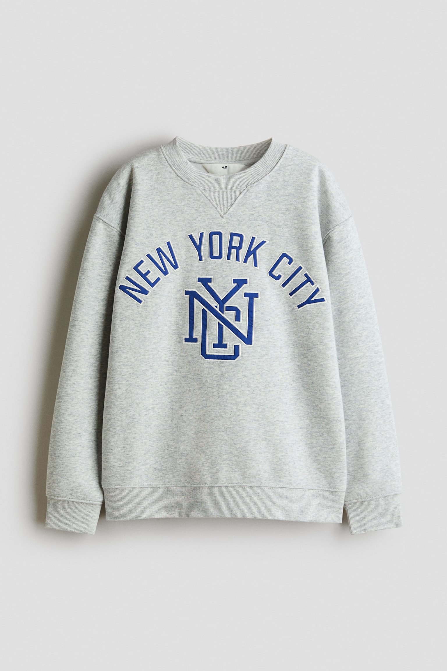 Má»t chiáº¿c Ã¡o sweatshirt cá» trÃ²n mÃ u xÃ¡m nháº¡t melange ÄÆ°á»£c chá»¥p tá»« phÃ­a trÆ°á»c, vá»i thiáº¿t káº¿ vai trá», bo tay Ã¡o gÃ¢n, lai Ã¡o gÃ¢n vÃ  máº£ng chÃ¨n hÃ¬nh tam giÃ¡c gÃ¢n á» cá» Ã¡o. Pháº§n ngá»±c in há»a tiáº¿t chá»¯ cong âNEW YORK CITYâ mÃ u xanh viá»n tráº¯ng, phÃ­a trÃªn lÃ  monogram âNYCâ lá»n Äan lá»ng vÃ o nhau.