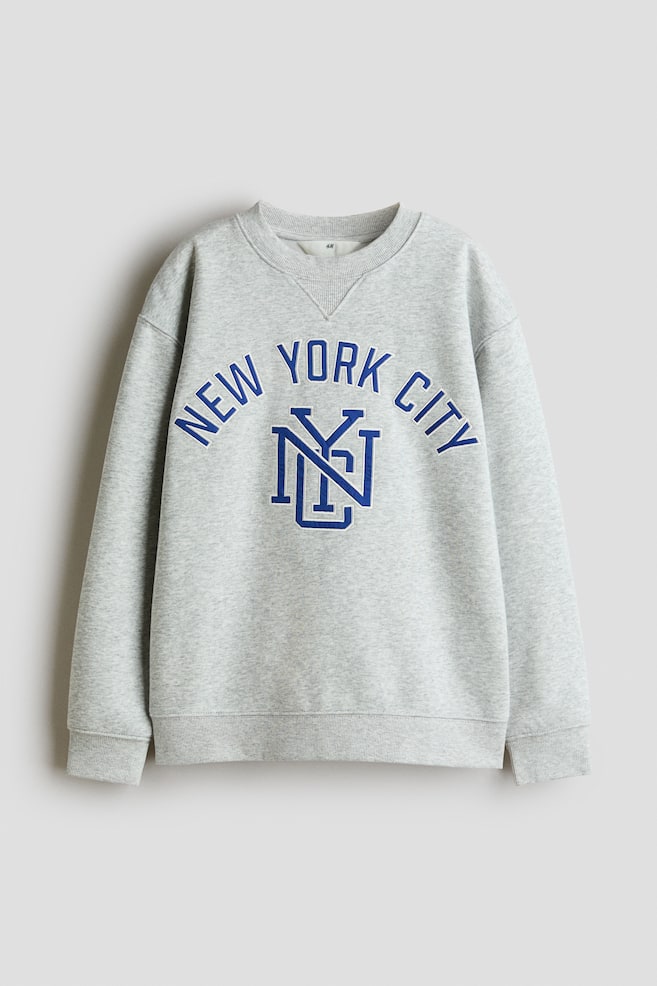 A light grey melange crewneck sweatshirt is shown from the front, featuring dropped shoulders, ribbed cuffs, a ribbed hem, and a ribbed triangle insert at the collar. The chest is displaying an arched blue "NEW YORK CITY" graphic with a white outline above a large, interlaced "NYC" monogram. - 1