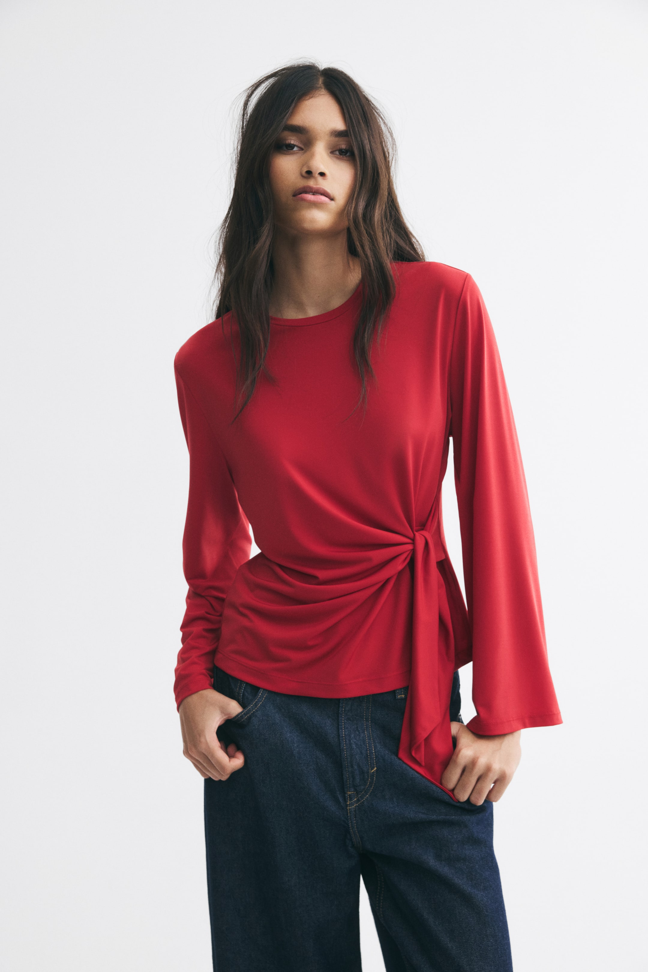 View larger image: The model is wearing a red long-sleeve stretch jersey top featuring a round neckline and a side-tie detail at the waist, creating a draped effect. The top is styled with dark blue jeans, with the model resting their hands in the front pockets.