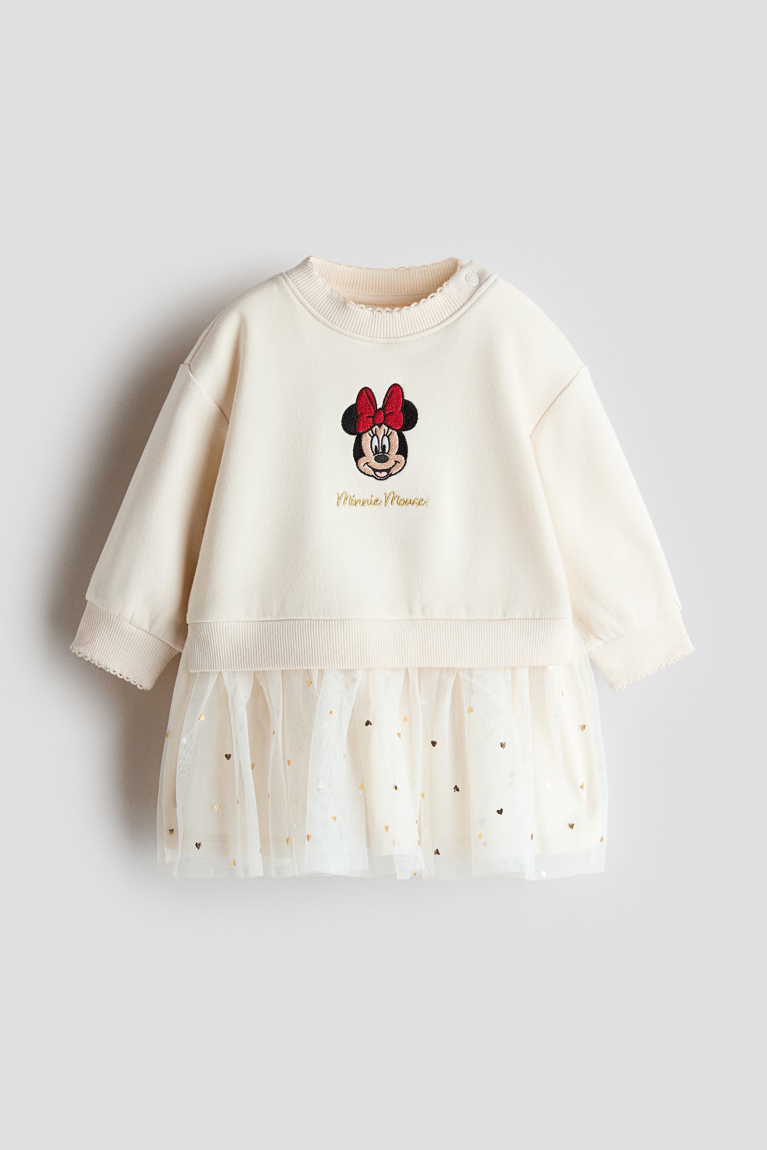 A cream-coloured sweatshirt dress showcases a front-facing design with an embroidered Minnie Mouse graphic and gold script on the chest. The dress features a ribbed crew neck and ribbed cuffs, while the lower half highlights a gathered white tulle skirt overlay decorated with small, shiny gold hearts.
