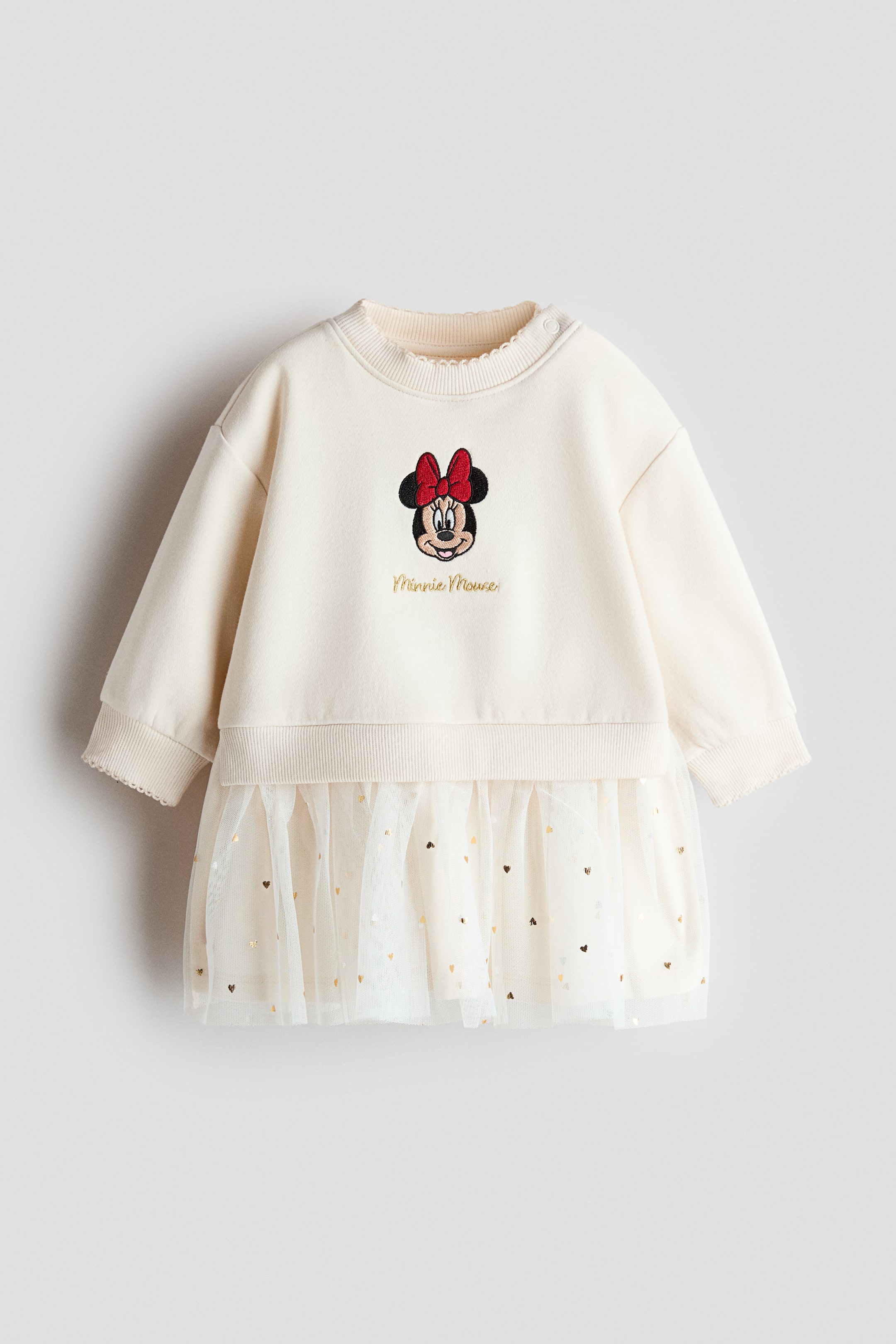 查看大圖: 一件米白色衛衣裙以正面展示,胸前飾有 Minnie Mouse 刺繡圖案及金色手寫體字樣。裙款採用羅紋圓領及羅紋袖口,下半部分為聚褶白色薄紗裙擺覆層,並點綴細小閃亮的金色愛心。