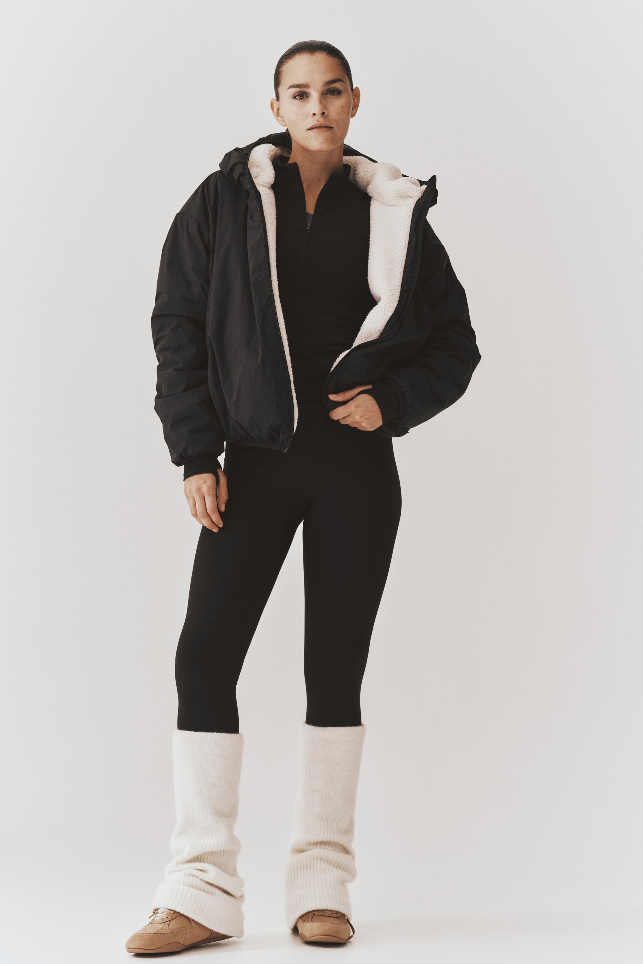 View larger image: An oversized, reversible activewear jacket with a black woven outer shell and a plush cream-colored teddy lining is worn open by the model. The jacket features a hood and dropped shoulders and is styled over a black mock neck top, black leggings, and slouchy cream leg warmers.