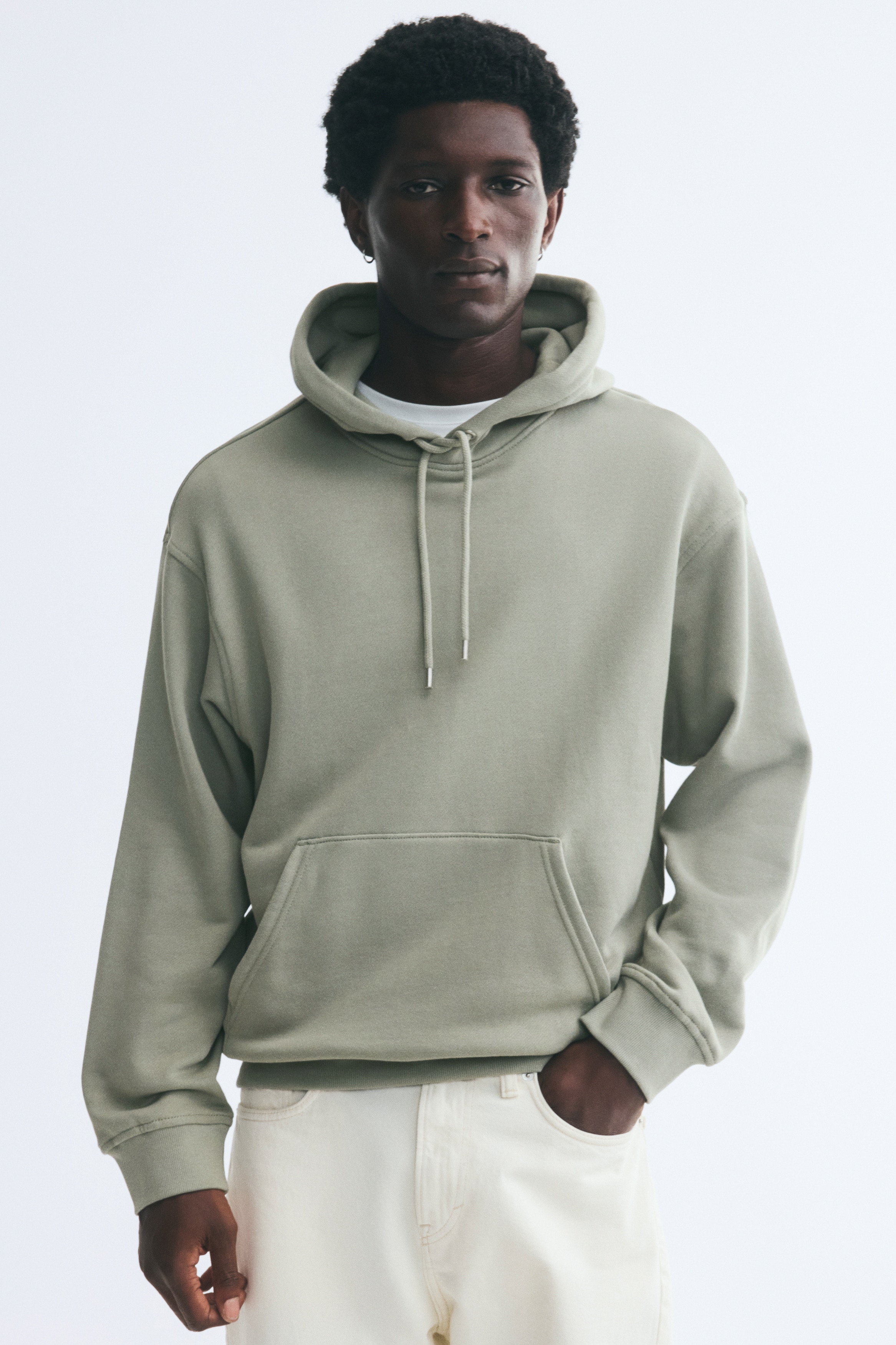 A light green hoodie is shown from the front, featuring a relaxed fit with dropped shoulders, long sleeves, and a jersey-lined drawstring hood. The garment showcases a front kangaroo pocket, along with ribbed cuffs and hem.