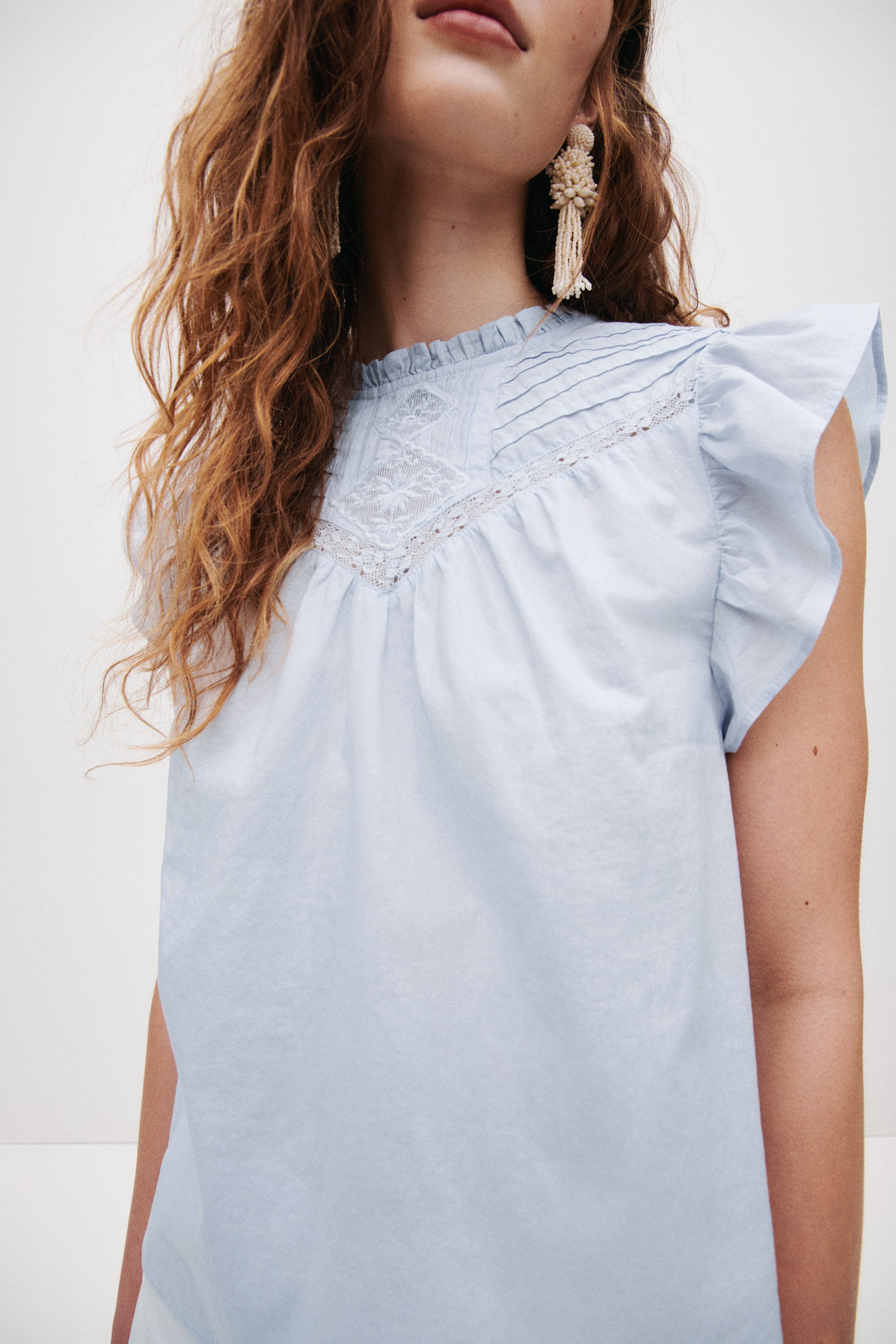Ingrandisci l'immagine: Un top azzurro chiaro con un collo alto arricciato, un pannello in pizzo a forma di V sul carré affiancato da pieghe e un delicato bordo in pizzo, e maniche corte arricciate.