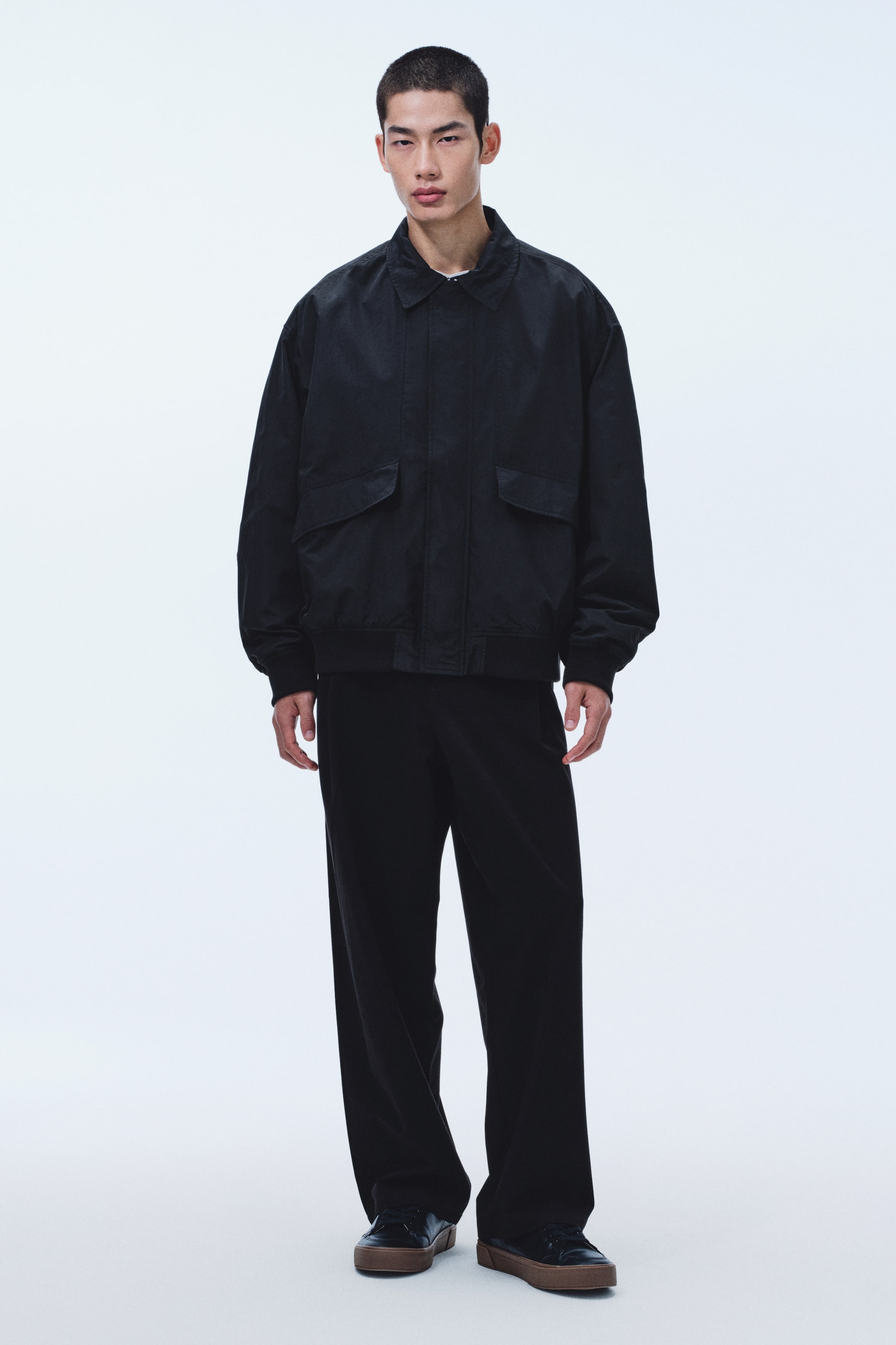 拡大画像を表示: ブラックのボンバージャケットは、ポインテッドカラー、フロントに大きなフラップポケットが2つ、リブカフスとリブヘムを備え、下に白いTシャツをレイヤードしてネックラインから見えています。