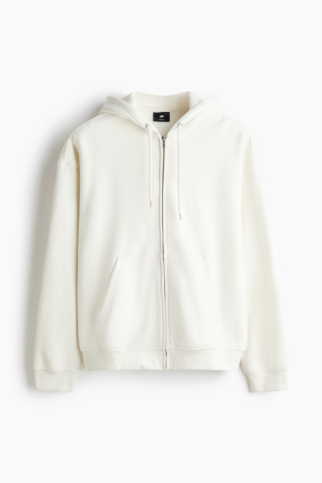 A cream-colored zip-through hoodie is being presented as FRONT, featuring a hood with a drawstring and a full-length front zipper. It is designed with dropped shoulders, long sleeves, and ribbed cuffs and hem, and a large kangaroo pocket is visible at the front, creating a loose and relaxed silhouette. - 1