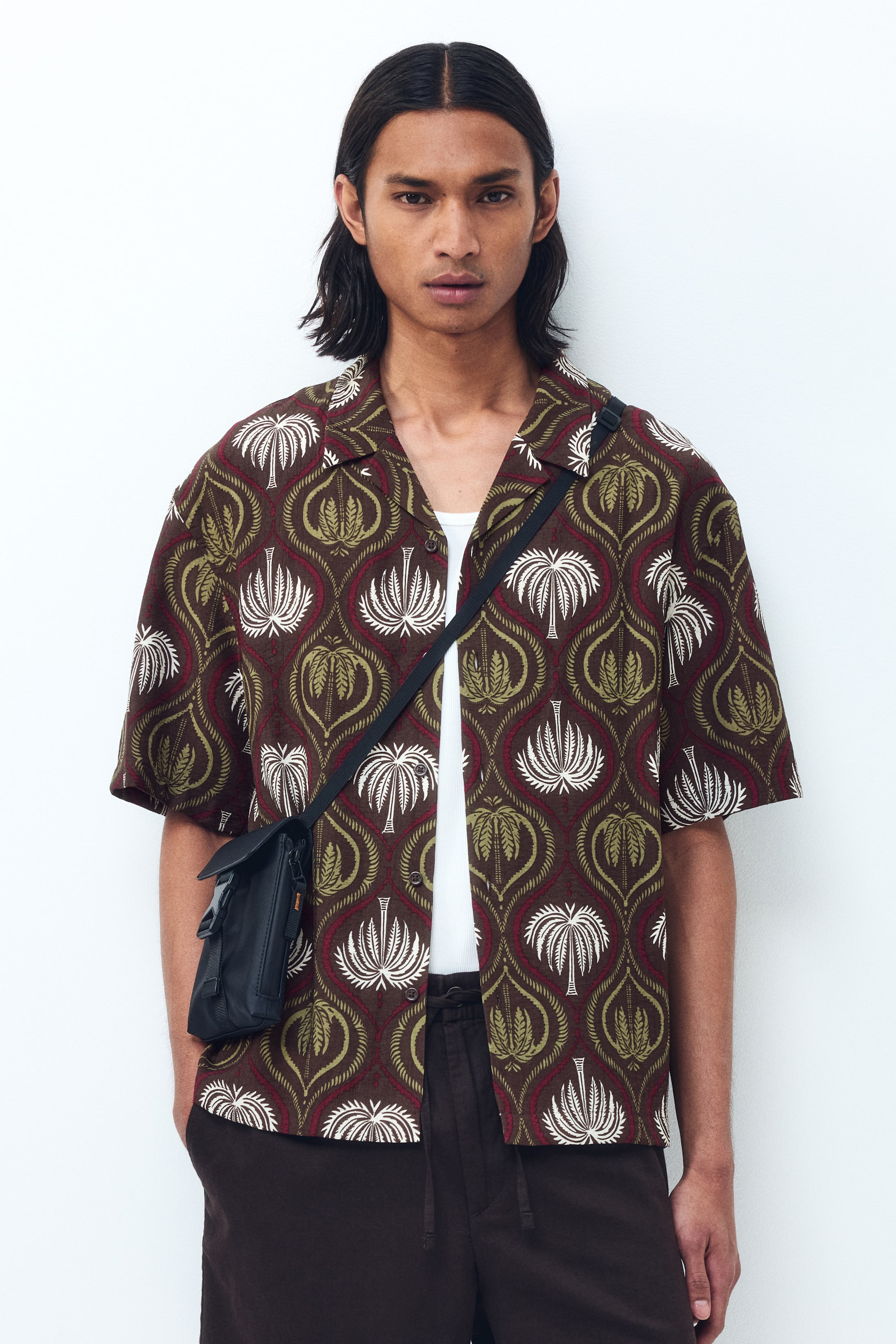 View larger image: "Model mengenakan baju kemeja berlengan pendek dengan anyaman bercorak yang ringan, menampilkan kolar resort, bahagian depan gaya Perancis, hem potongan lurus, dan potongan santai. Kemeja ini digayakan dengan butang terbuka di atas baju dalam putih, dan beg silang hitam dipakai di atas kemeja."