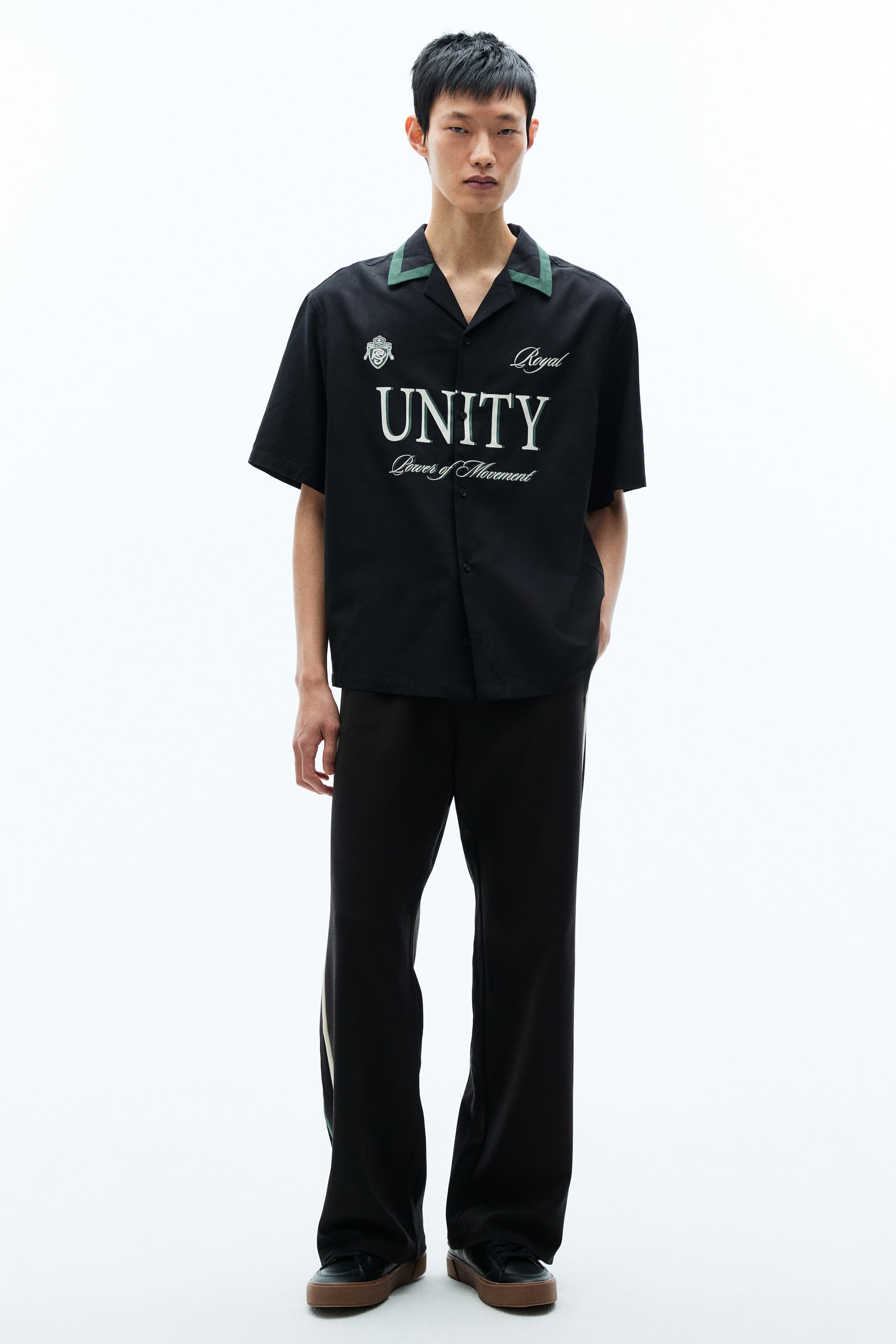 View larger image: A black short-sleeved shirt and black wide-leg trousers are being presented. The shirt features a green-trimmed revere collar, with 'Royal' in script, 'UNITY' in large capitals, and 'Power of Movement' in smaller script on the front, alongside a crest emblem. The trousers feature a light-coloured vertical stripe with a green accent along the side seams.