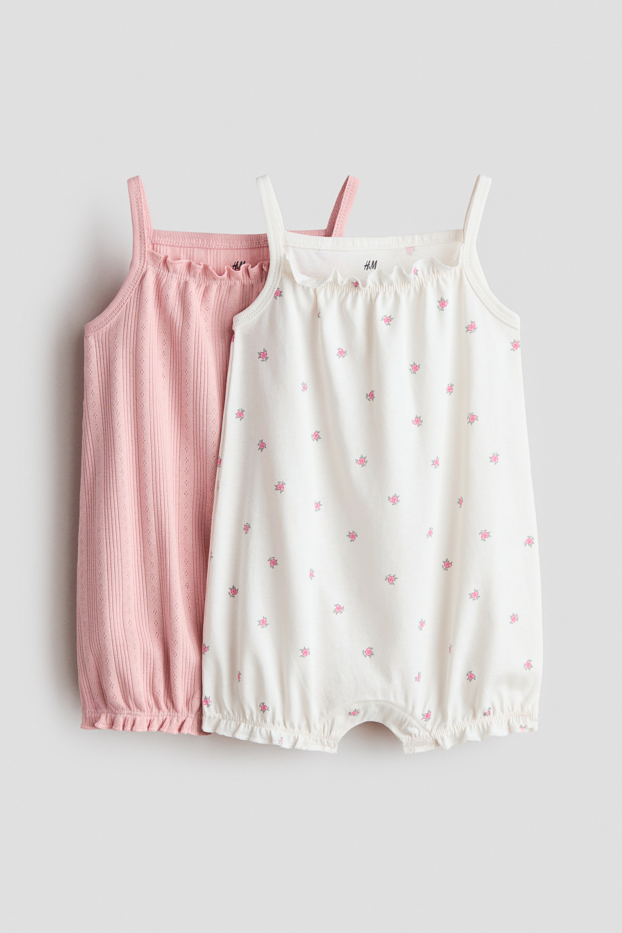 Agrandir l'image: Deux combinaisons pour bébé sont présentées, avec la combinaison blanche orientée vers l'avant. La combinaison blanche arbore un petit motif floral rose, des bretelles fines, un col carré élastiqué à volants et des ouvertures pour les jambes à volants, tandis que la combinaison rose clair, apparaissant derrière, présente un tricot ajouré côtelé avec des bretelles fines et un col et un ourlet inférieur élastiqués à volants.