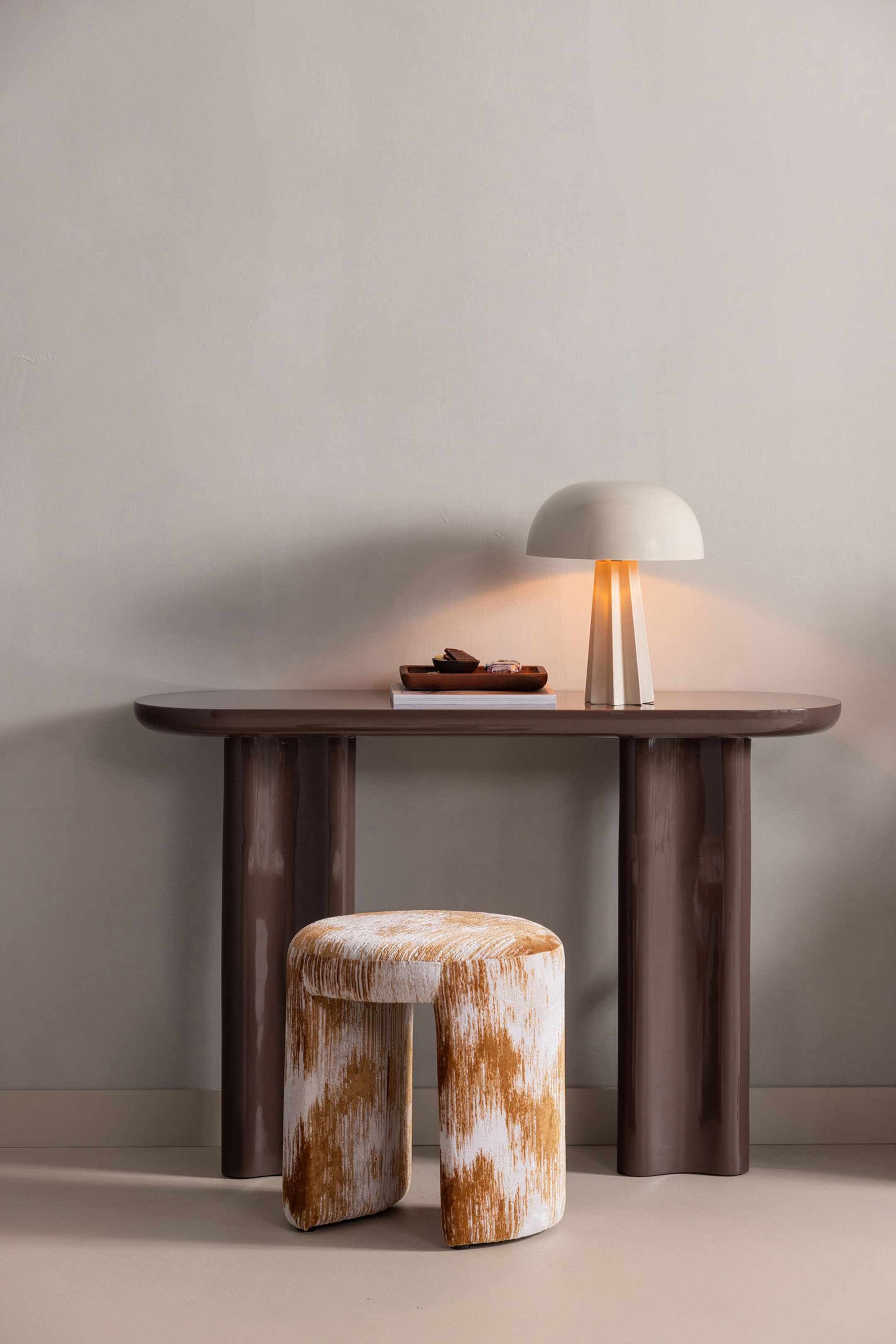 Agrandir l'image: Une table console marron foncé est orientée vers l'avant, avec un plateau épais et arrondi et deux larges pieds incurvés. Un tabouret en forme d'arc avec un motif abstrait marron et blanc est placé sous la table, et une lampe de couleur crème repose sur sa surface.
