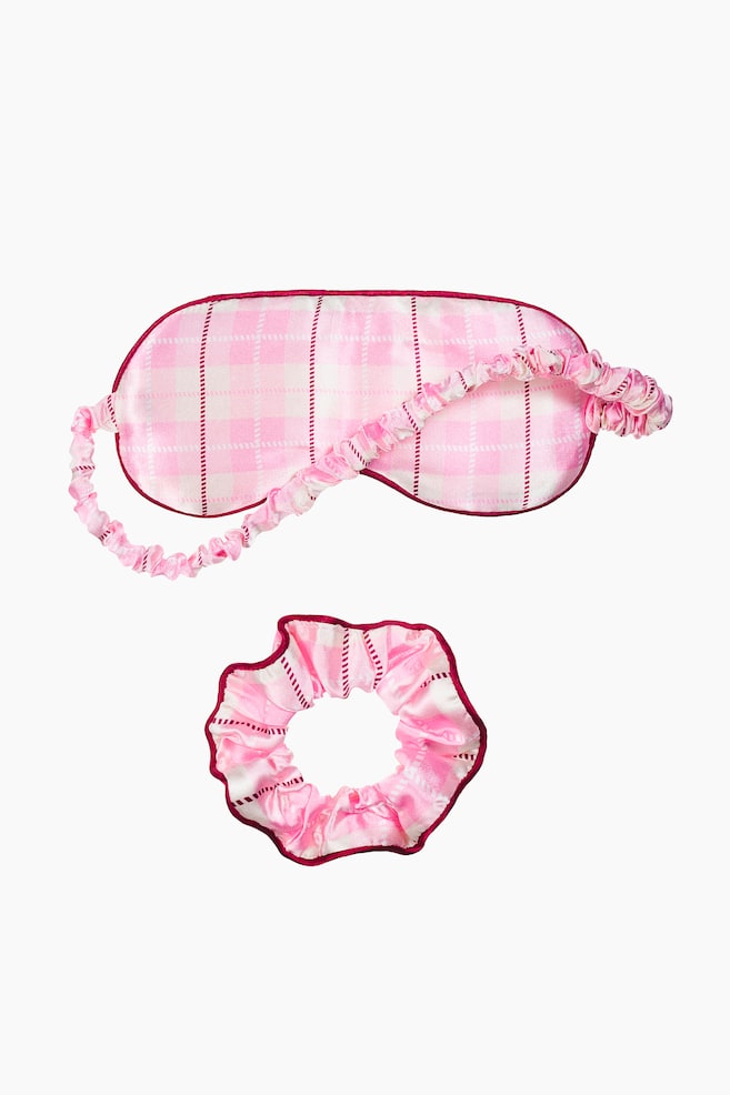 A pink and white plaid satin sleep set is displaying a matching sleep mask and scrunchie from the front. The sleep mask is featuring a padded eye cover with burgundy piped edges and a gathered elastic strap, while the coordinating scrunchie is showing a ruffled design with a matching burgundy trim. - 1