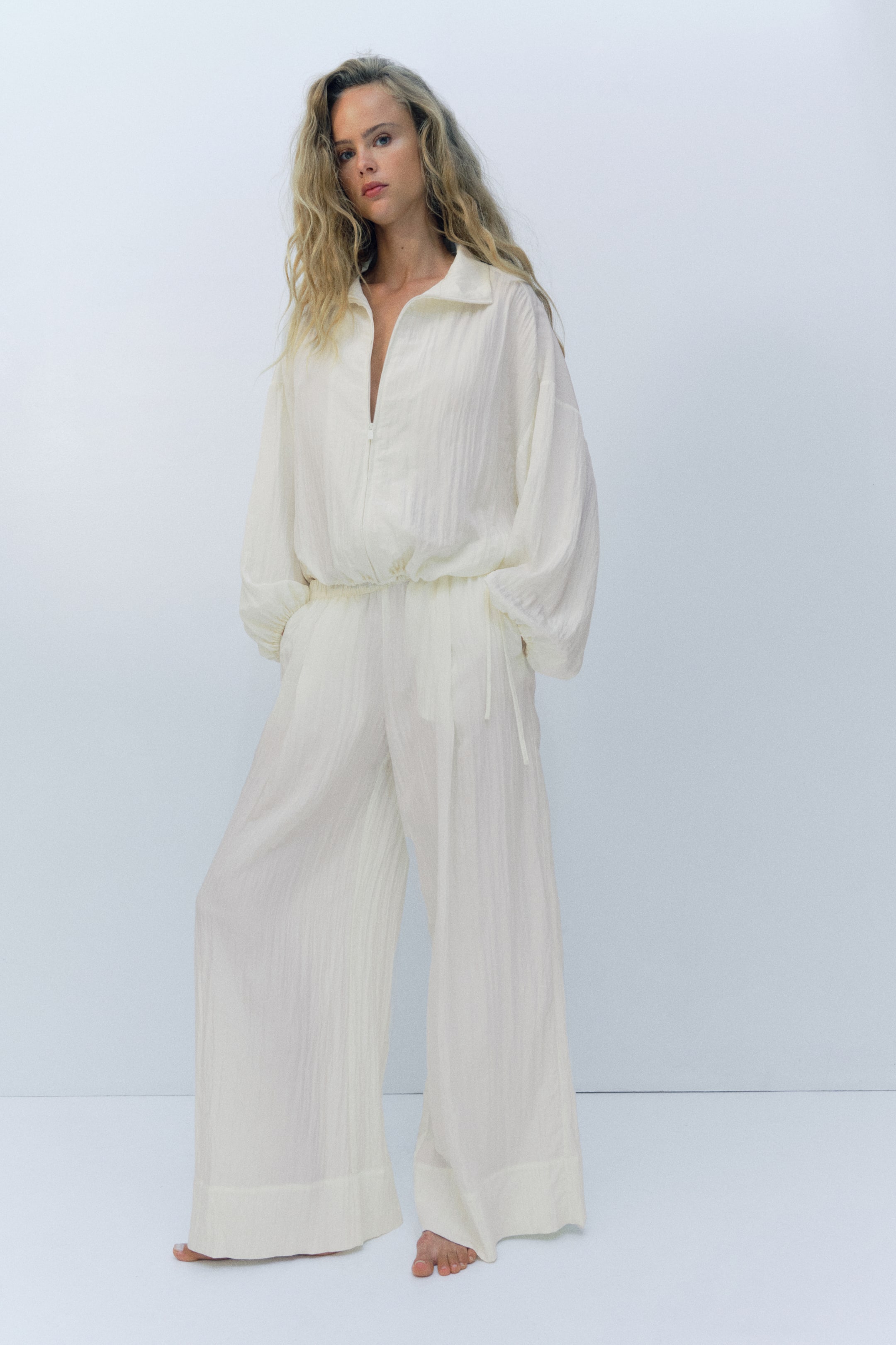 Größeres Bild ansehen: Die Model trägt eine locker geschnittene, weit geschnittene Hose aus gekräuseltem, cremeweißem Stoff, kombiniert mit einem passenden Langarm-Top mit Reißverschluss. Die Hose verfügt über einen elastischen Bund und schräge Seitentaschen, in die das Model seine Hände steckt.