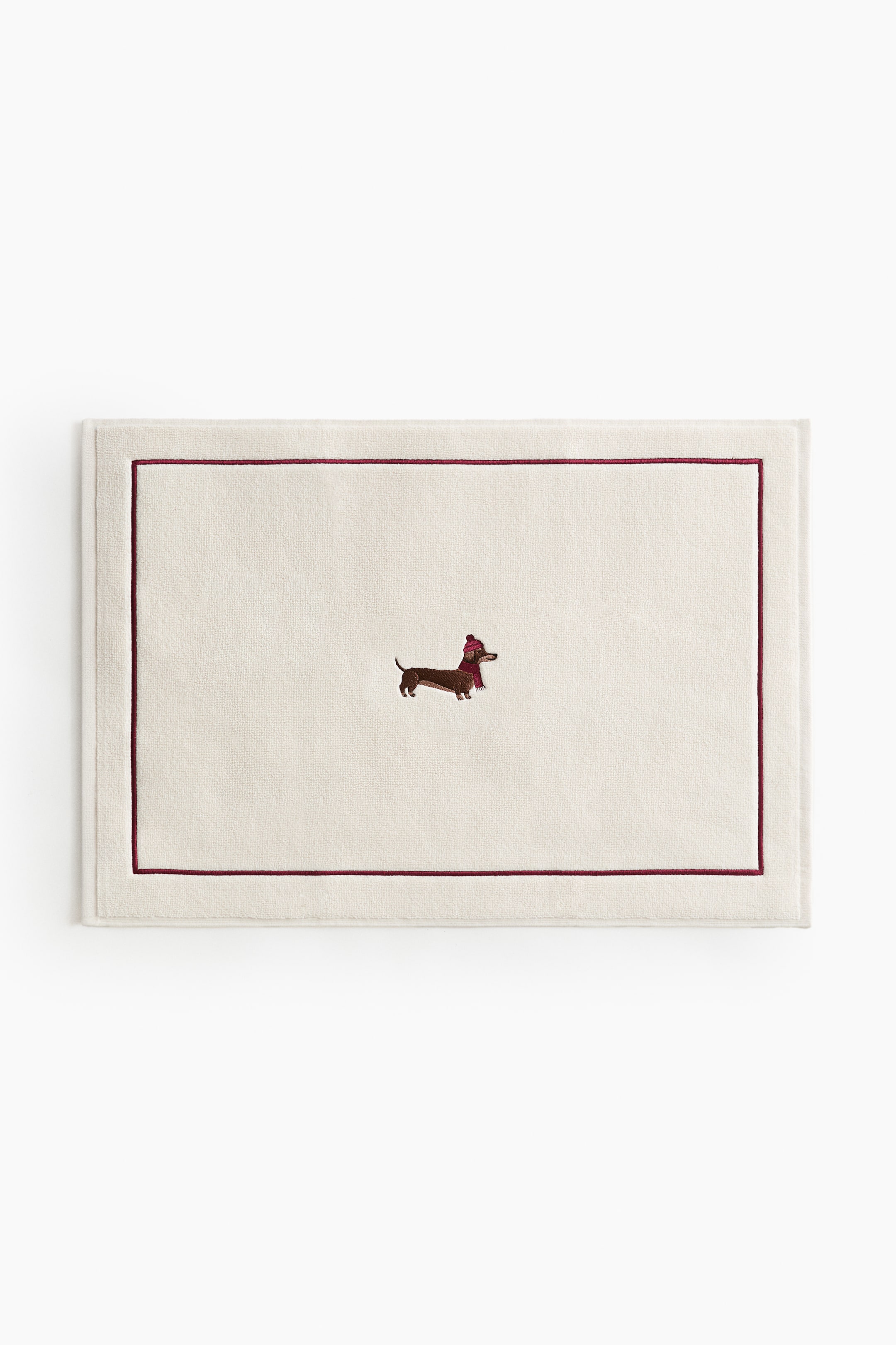 View larger image: A rectangular, off-white bath mat is shown from the front, featuring a thin burgundy embroidered border and a small embroidered dachshund wearing a red winter hat and scarf at the centre.