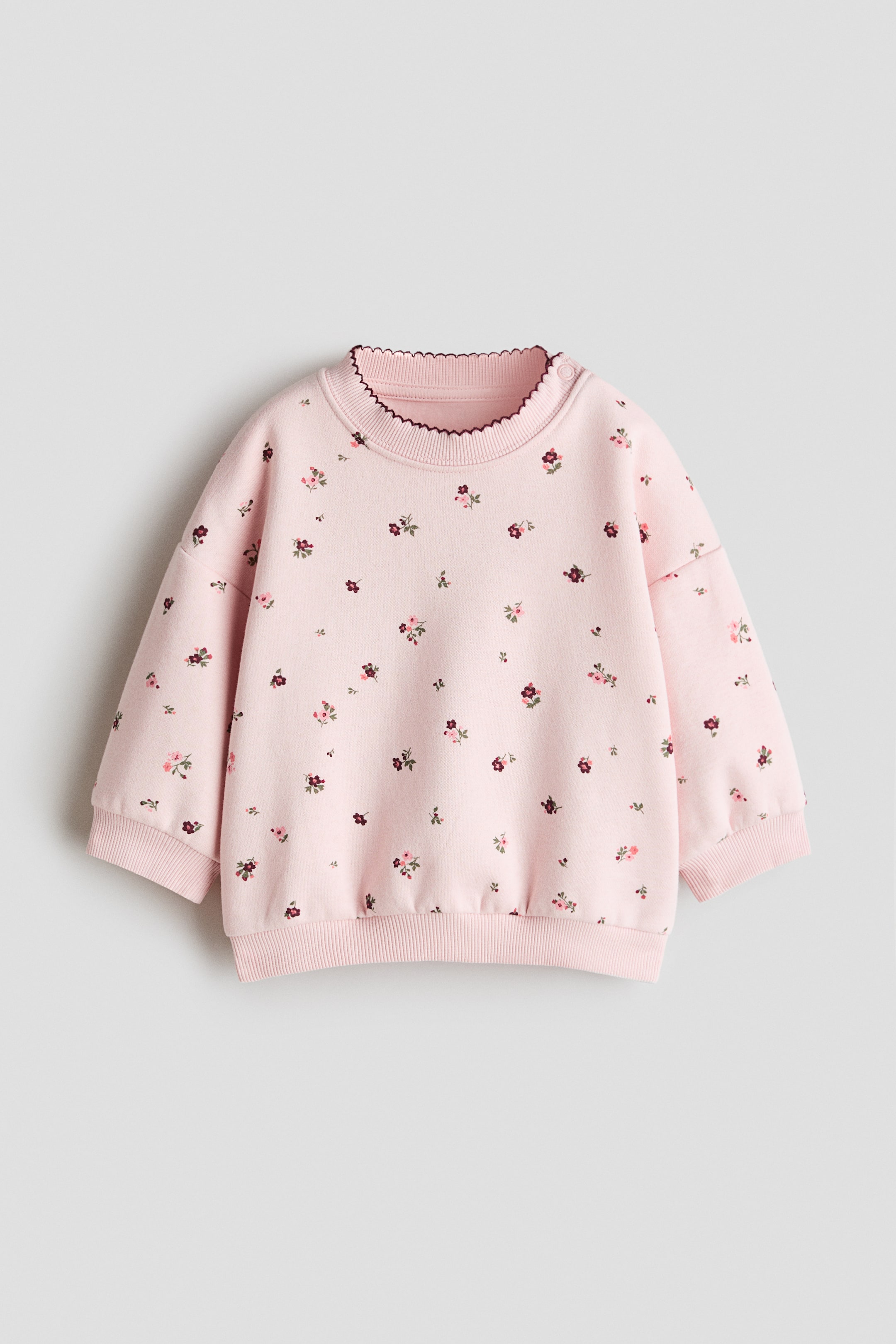 View larger image: A printed sweatshirt is laid flat, highlighting a light pink base adorned with a scattered floral pattern featuring small pink, red, and maroon flowers. It comes with a crew neck accentuated by a dark scalloped trim, two snap buttons on the left shoulder, and ribbed cuffs and hem.