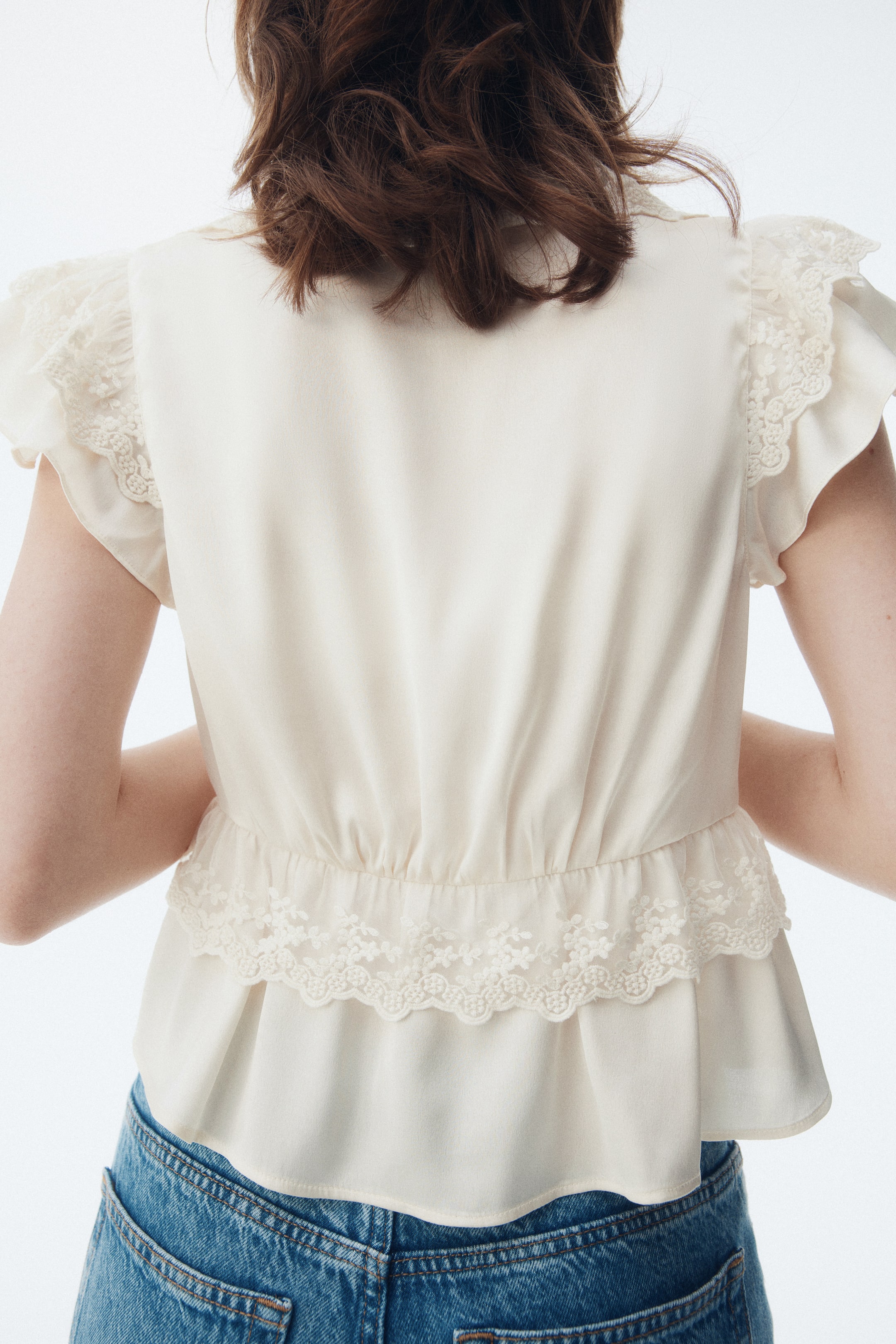 View larger image: A cream-colored top is being shown from the back, featuring short flutter sleeves that are trimmed with lace. The top is gathering at the waist, creating a peplum hem that is also edged with lace.