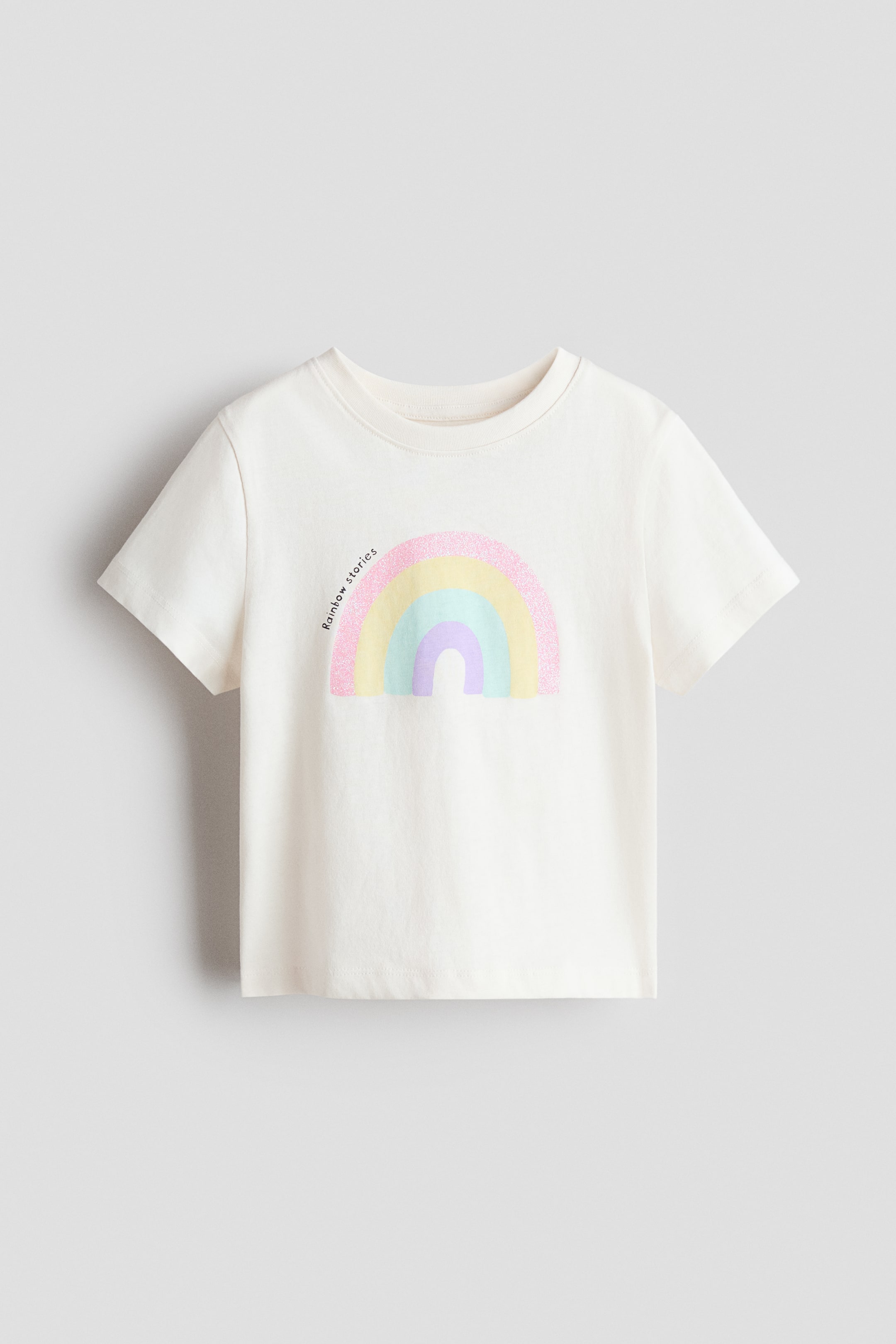 View larger image: A front-facing cream-colored T-shirt features a ribbed crew neck and short sleeves. The front displays a pastel rainbow graphic with a glittery pink outermost band, followed by yellow, light blue, and light purple, and the text 'Rainbow stories' is printed vertically alongside it.