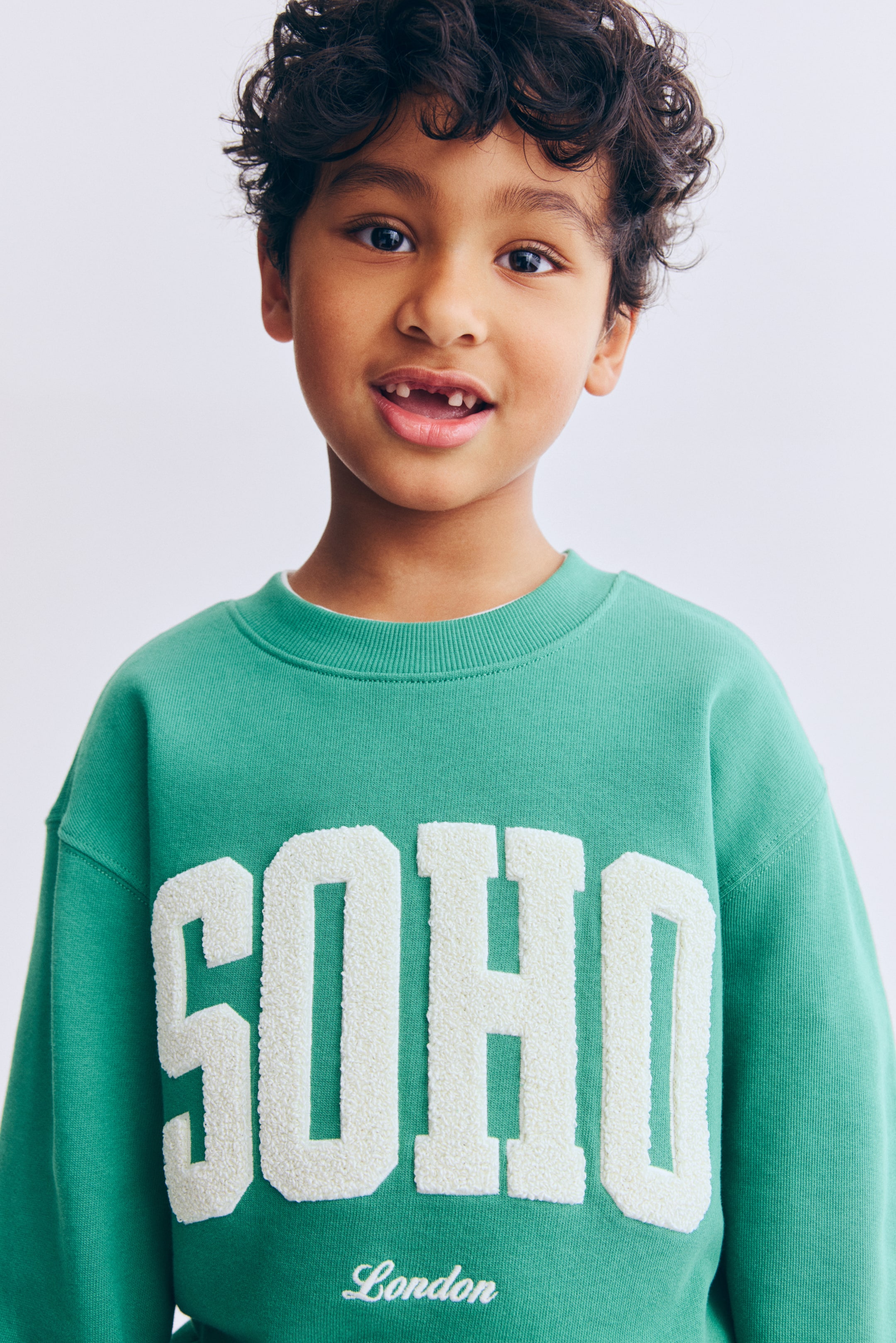 Zobacz większy obraz: Zielona bluza zaprezentowana od PRZODU, z okrągłym, prążkowanym dekoltem, obniżoną linią ramion oraz prążkowanymi ściągaczami przy mankietach i u dołu. Z przodu duży, fakturowany napis „SOHO” w złamanej bieli, z haftowanym poniżej na biało napisem „London”.